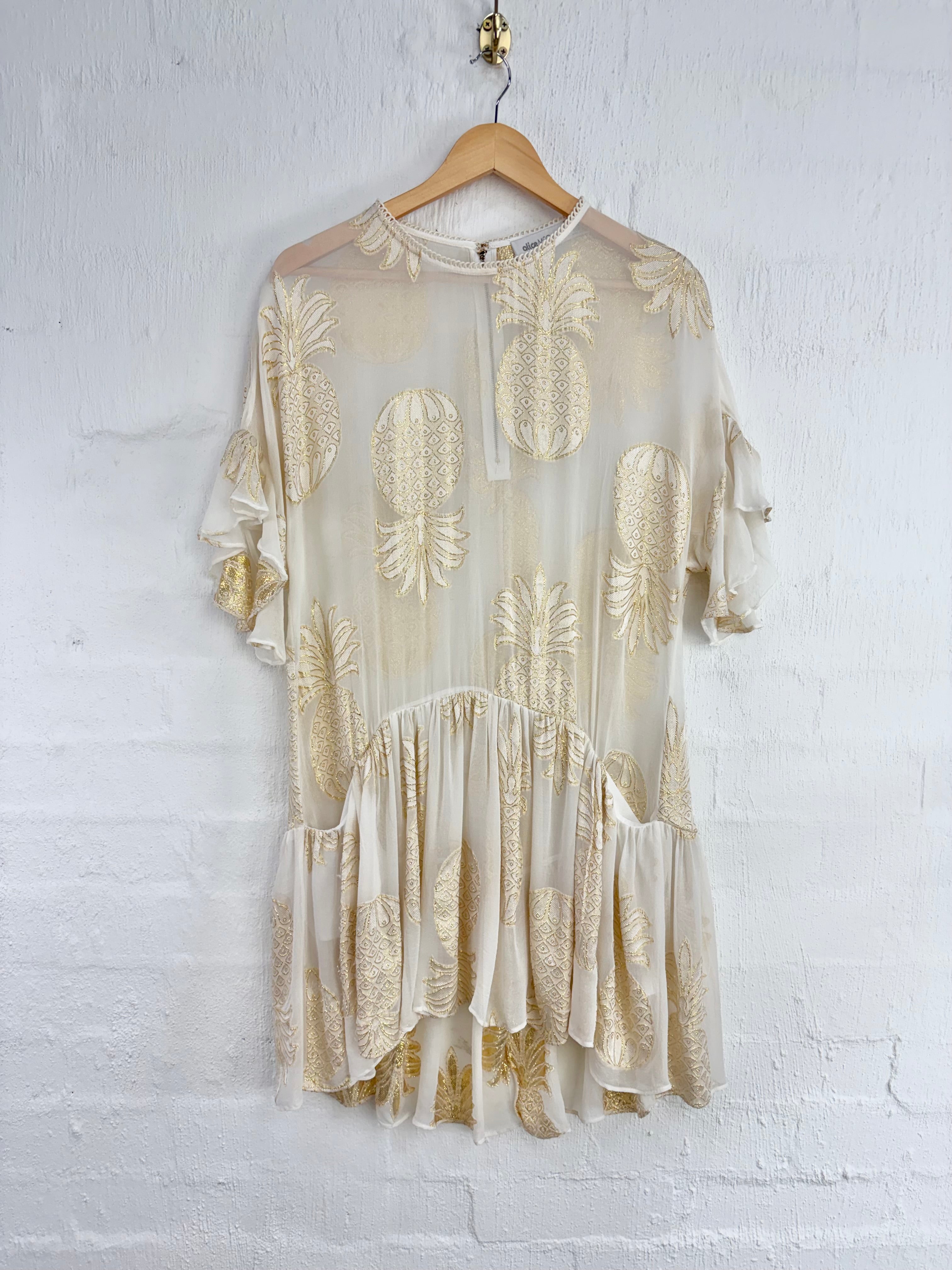 A cream-colored sheer mesh dress with gold pineapple embroidery, hanging on a wooden hanger against a white painted brick wall.