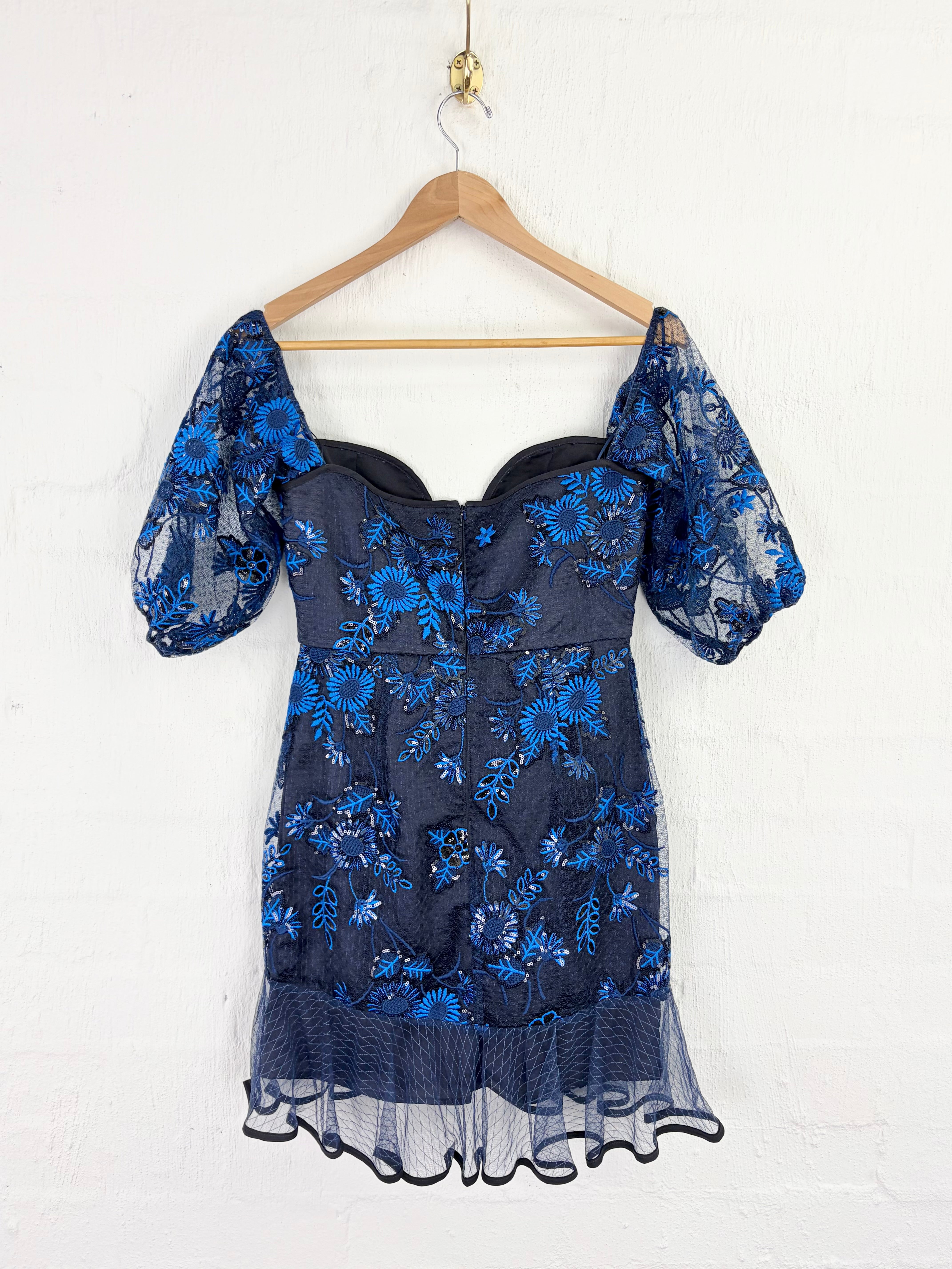 A dark navy blue lace dress with bright blue floral embroidery, featuring puff sleeves and a sweetheart neckline, hanging on a wooden hanger against a white brick wall.