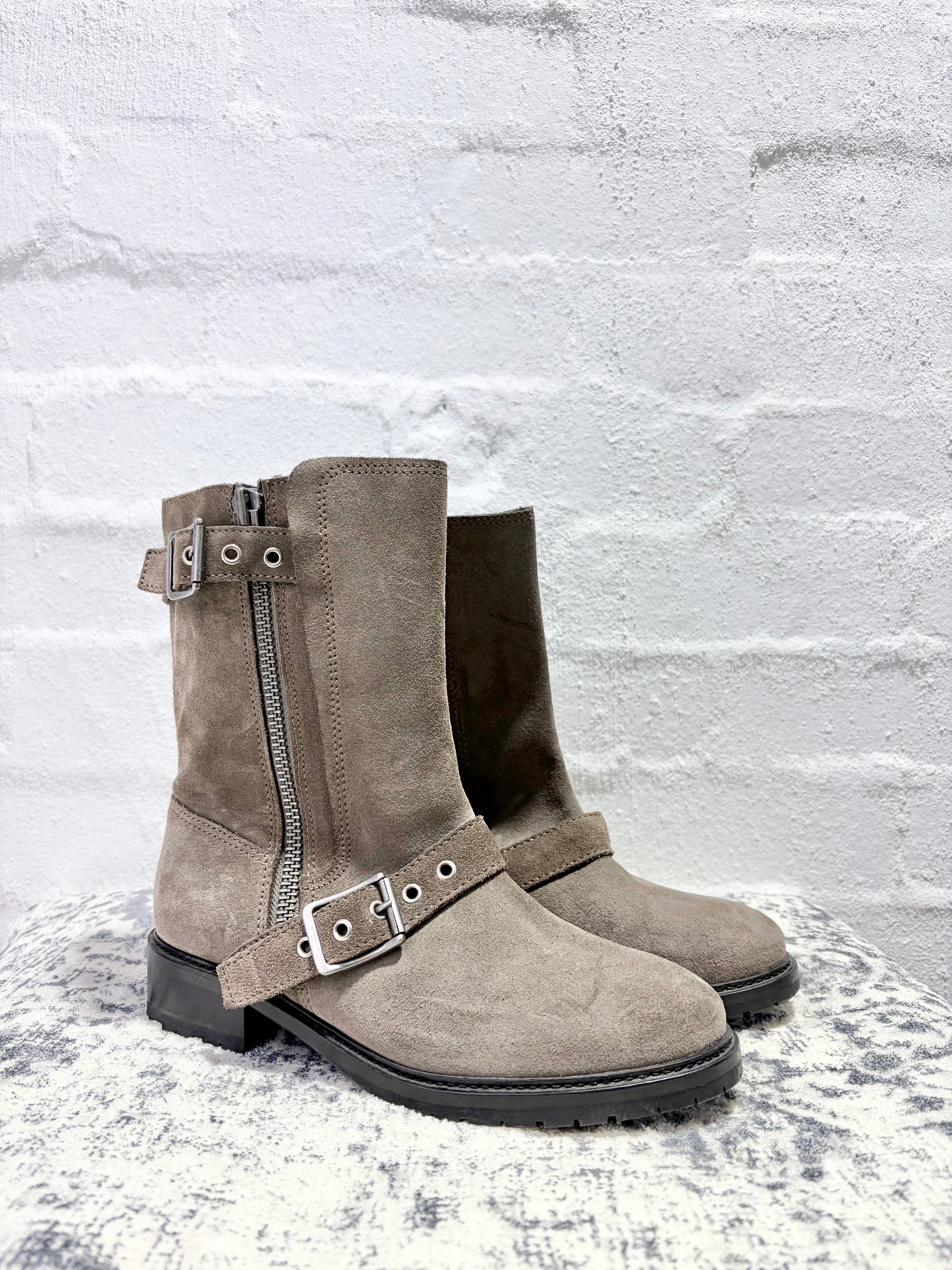 A pair of taupe-brown suede biker boots with side zippers and buckle straps, displayed on a textured surface against a white brick wall.