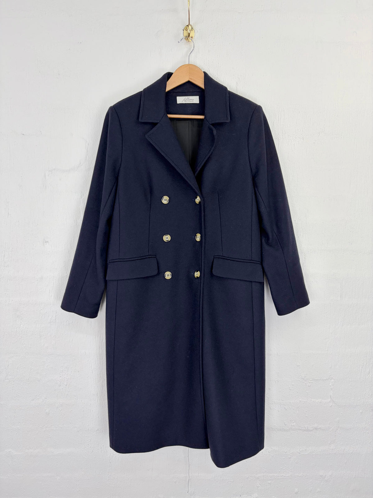 A navy blue double-breasted coat with gold buttons, hanging on a wooden hanger against a white painted brick wall.