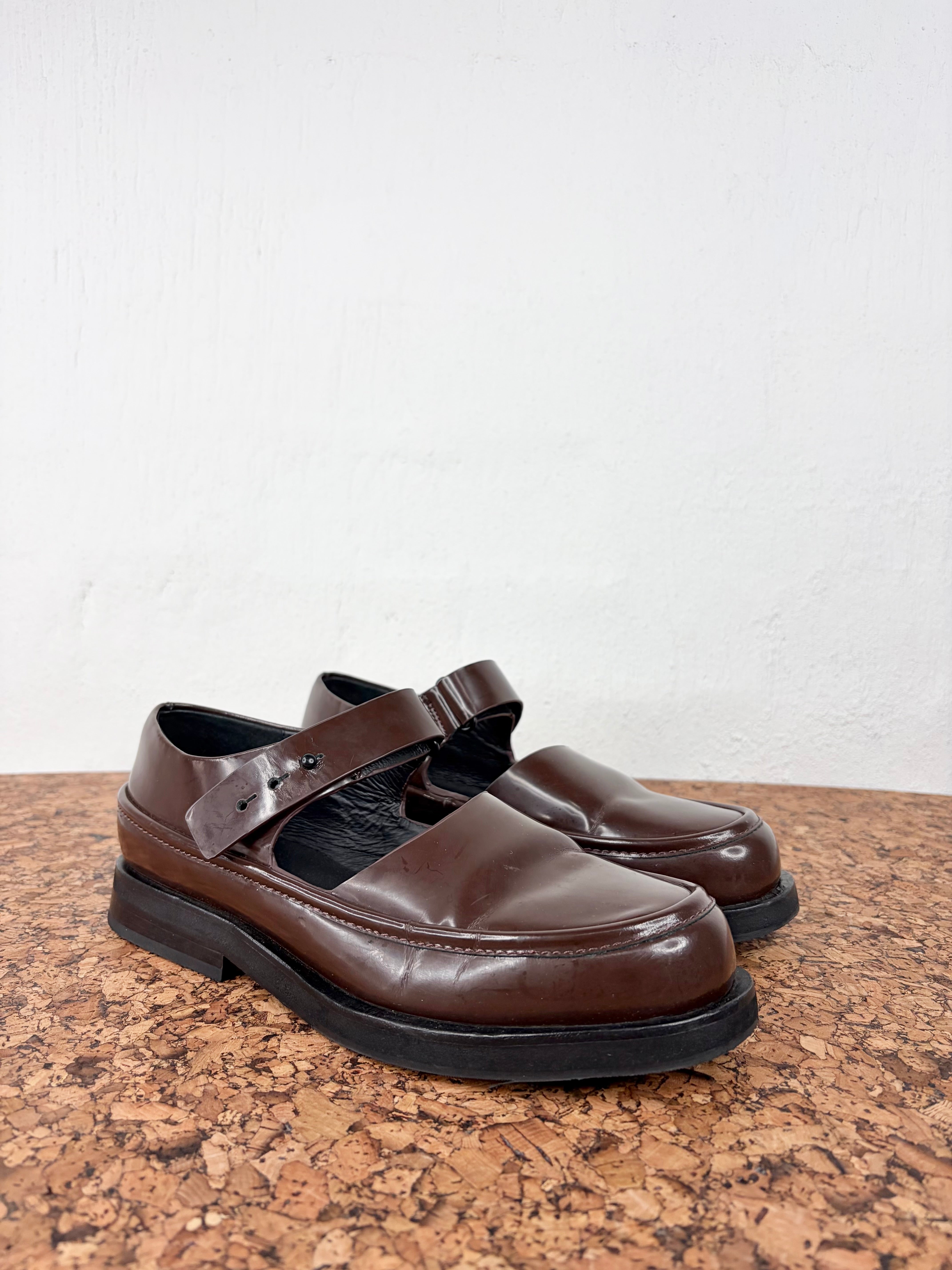 A pair of dark brown leather Mary Jane shoes with buckle straps, displayed on a cork surface against a white wall.