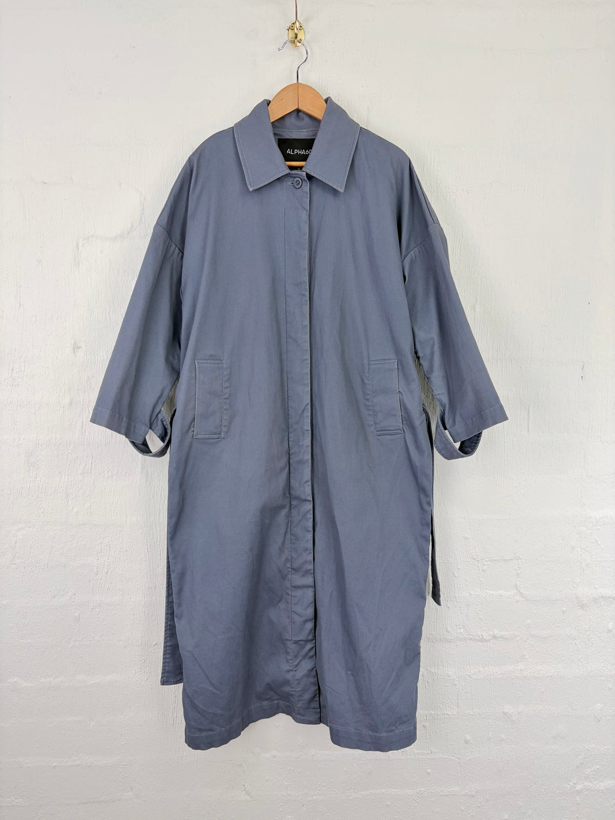 A slate blue button-up shirt dress with a pointed collar and three-quarter sleeves, hanging on a wooden hanger against a white painted brick wall.