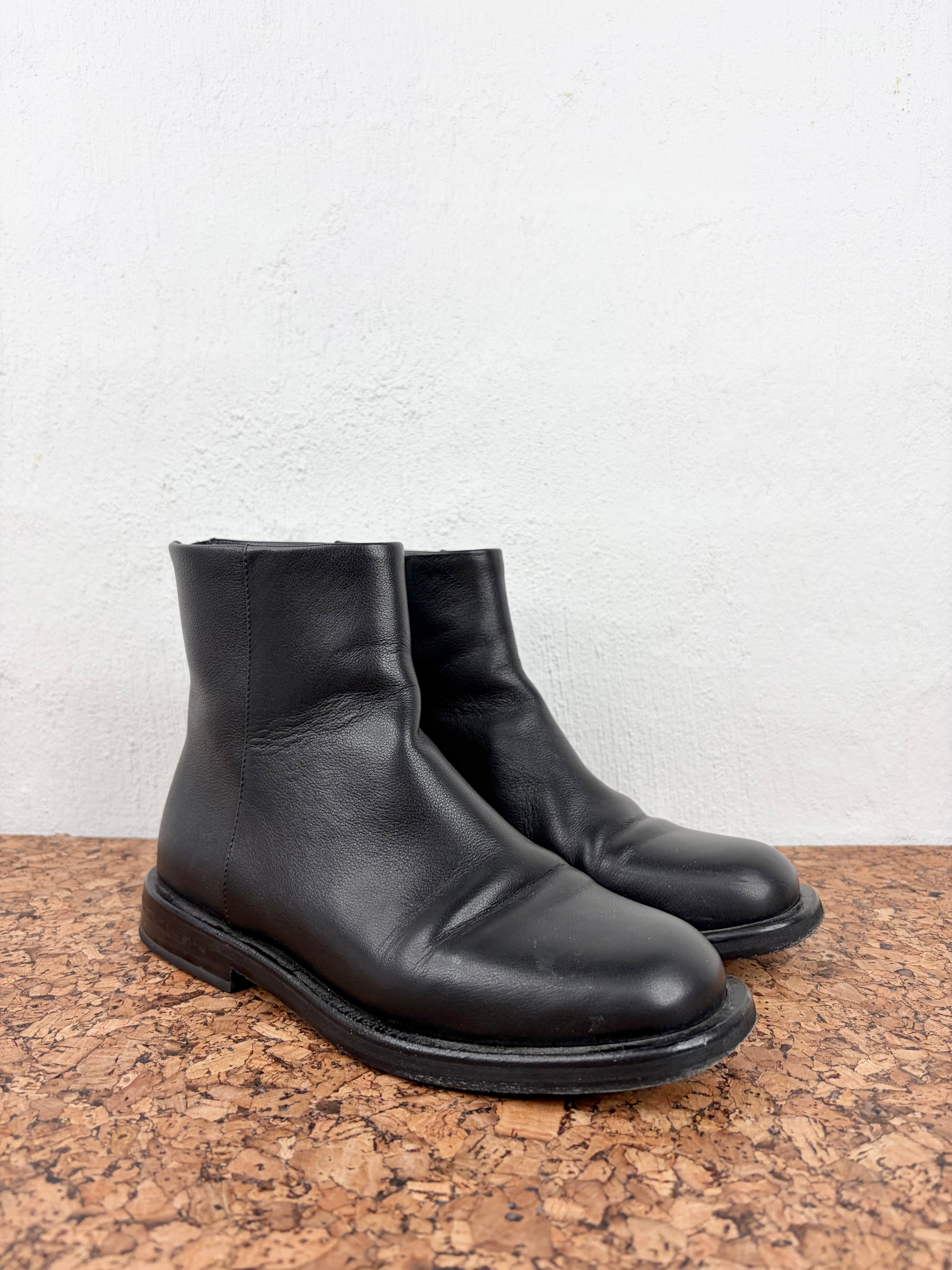 A pair of black leather ankle boots with a rounded toe and low stacked heel, resting on a cork surface against a white textured wall.