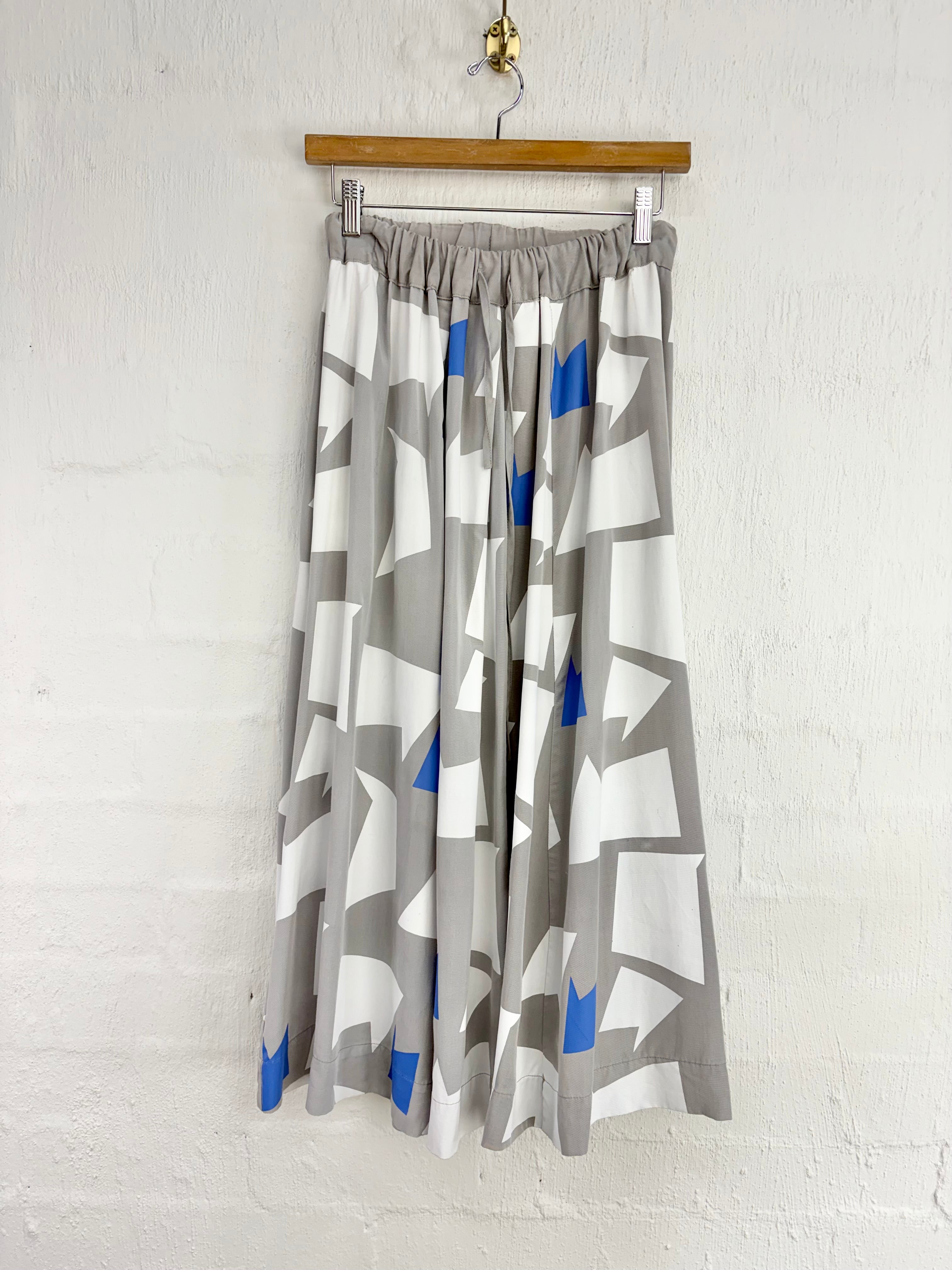 A pleated midi skirt with an abstract geometric pattern in gray, white, and blue, hanging from a wooden hanger against a white brick wall.