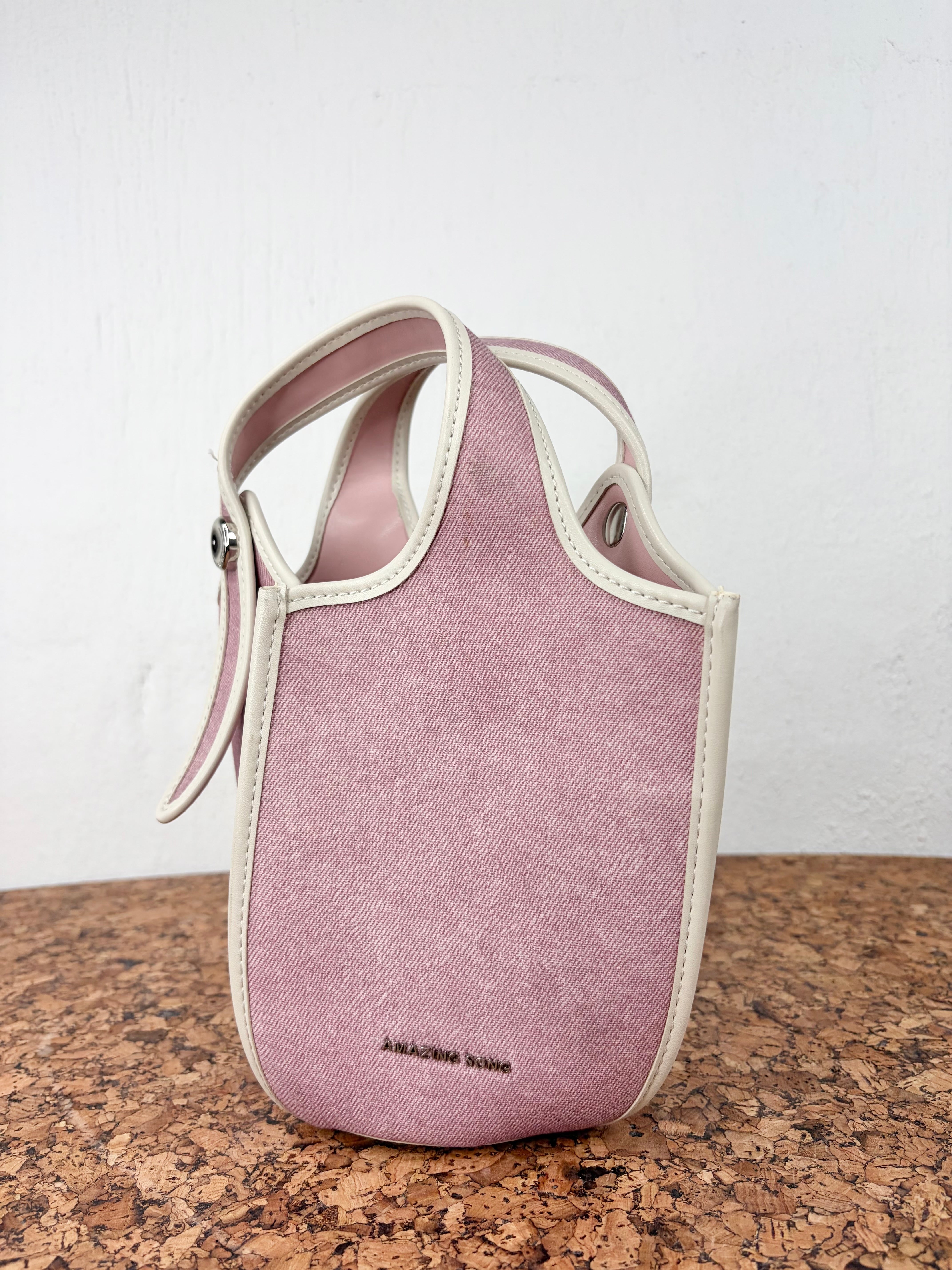 A small structured handbag in dusty pink canvas with cream-colored trim and a matching shoulder strap, standing on a brown cork surface against a white wall.