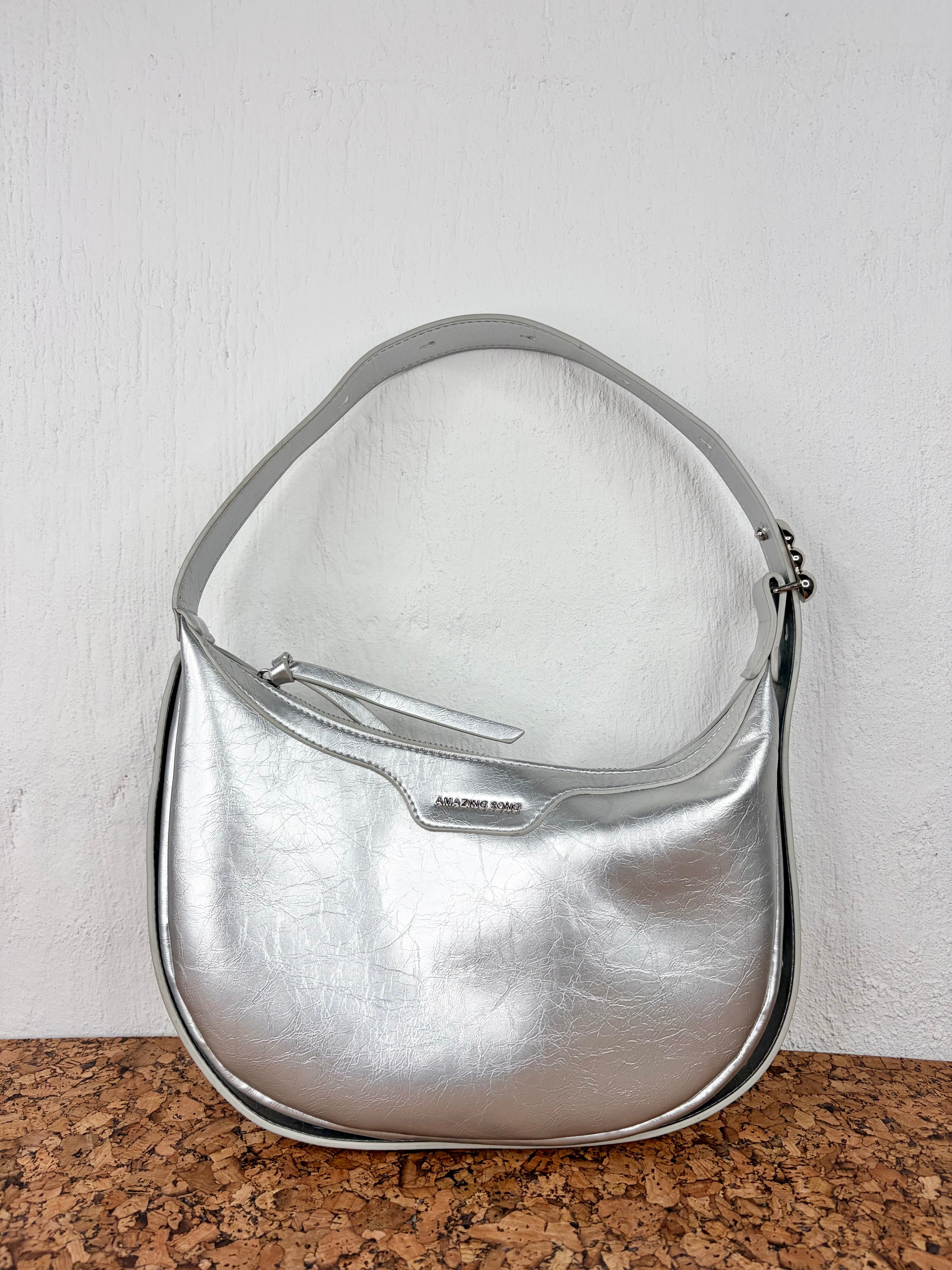 A metallic silver crescent-shaped shoulder bag with a zippered top and a small silver-tone logo plate, displayed on a brown cork surface against a white wall.