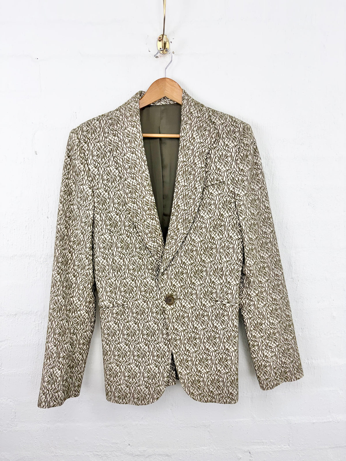 A single-breasted blazer jacket with a textured beige and brown houndstooth pattern, hanging on a wooden hanger against a white painted brick wall.