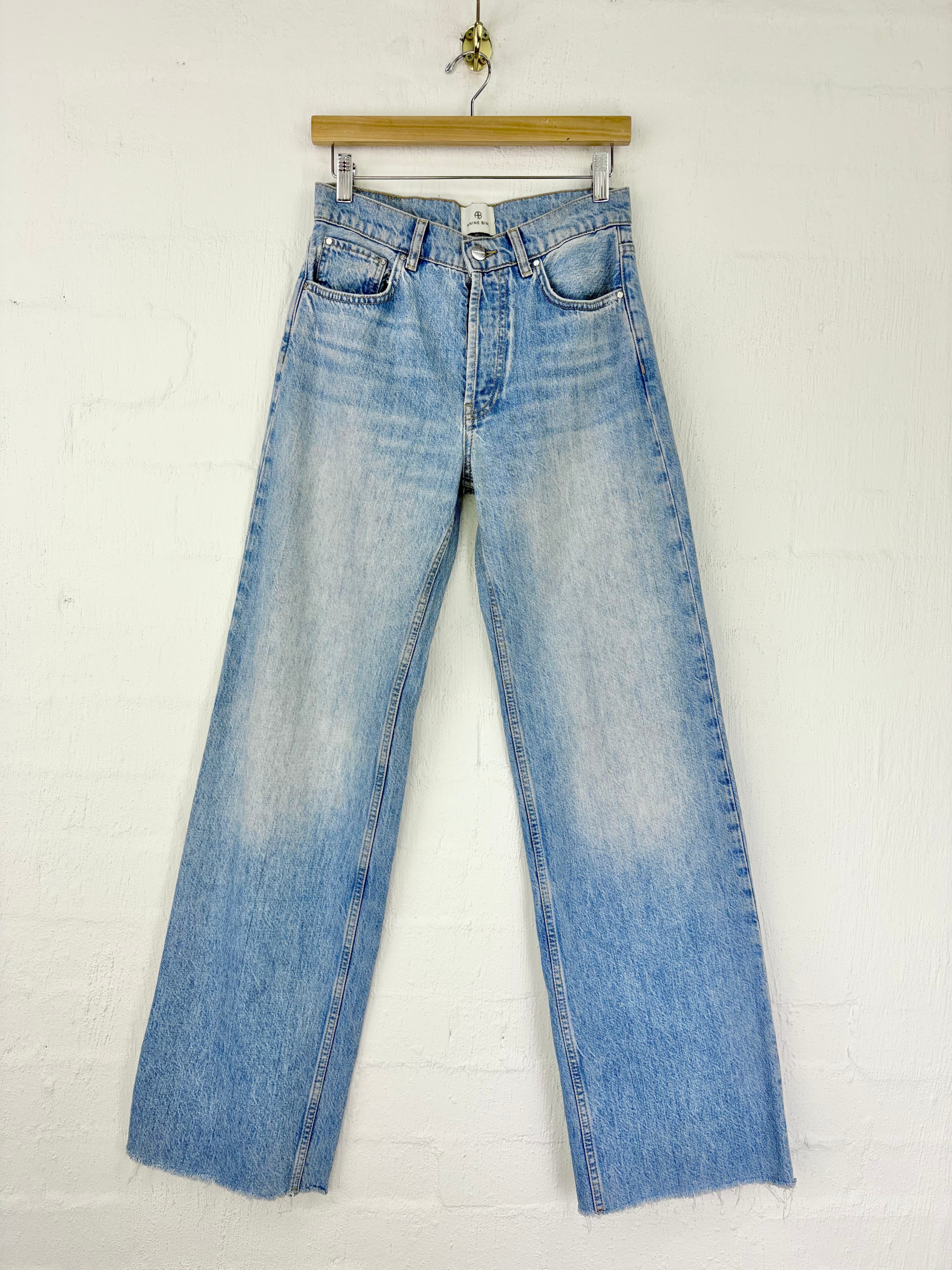 Light blue high-waisted straight-leg denim jeans with a faded wash, hanging from a wooden hanger against a white painted brick wall.