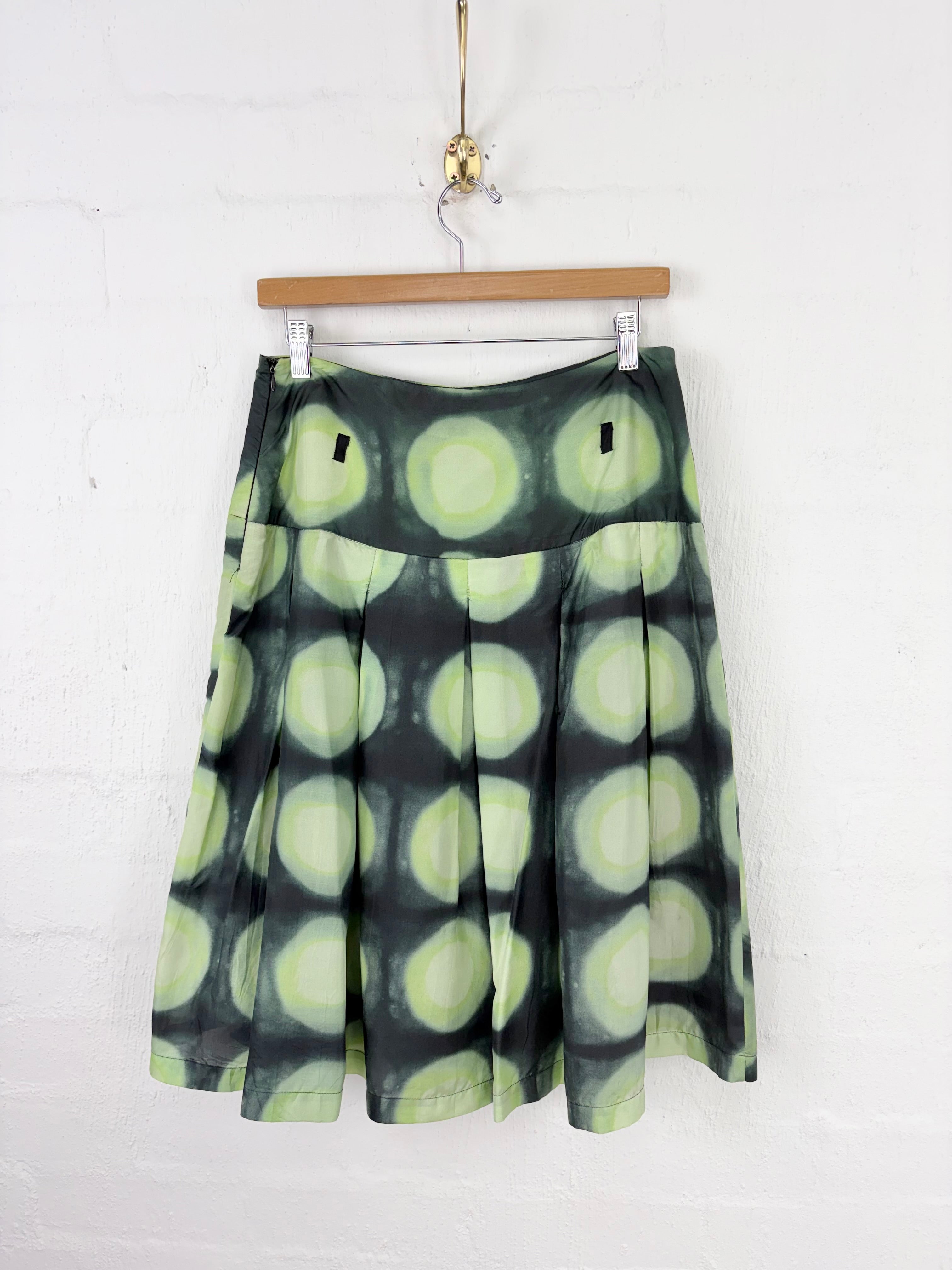 A pleated A-line skirt with a green and black bubble pattern, hanging on a wooden hanger against a white brick wall.