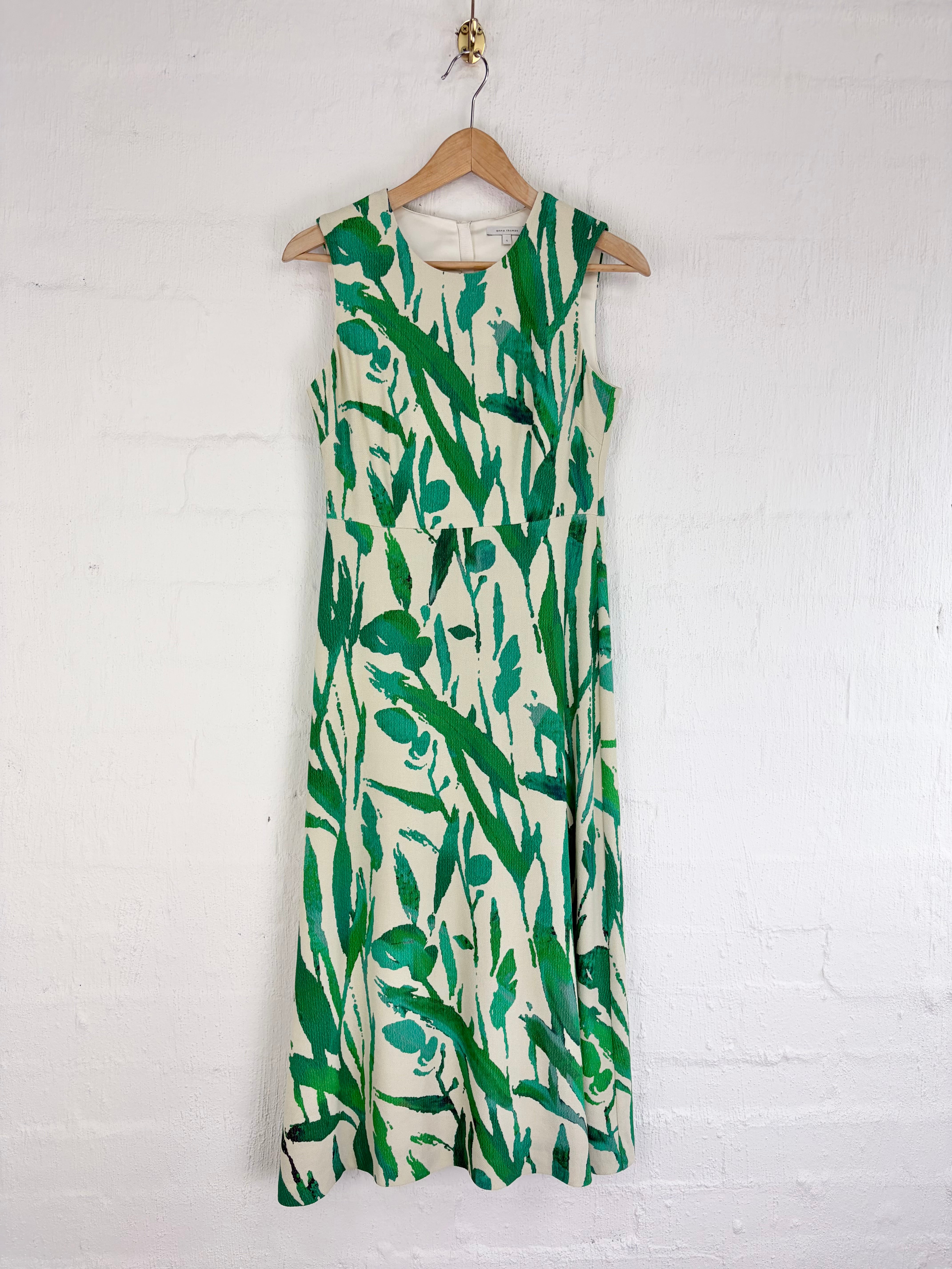 A sleeveless midi dress with a green botanical leaf print on a cream background, hanging on a wooden hanger against a white painted brick wall.