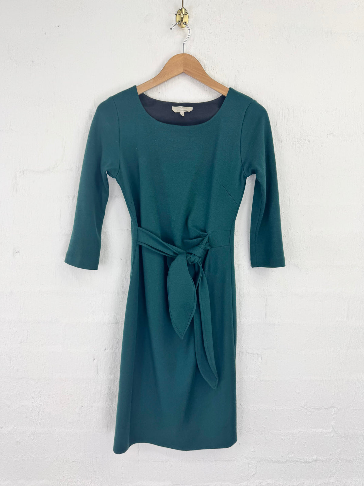 A teal green knee-length dress with three-quarter sleeves and a matching fabric tie belt at the waist, hanging on a wooden hanger against a white painted brick wall.