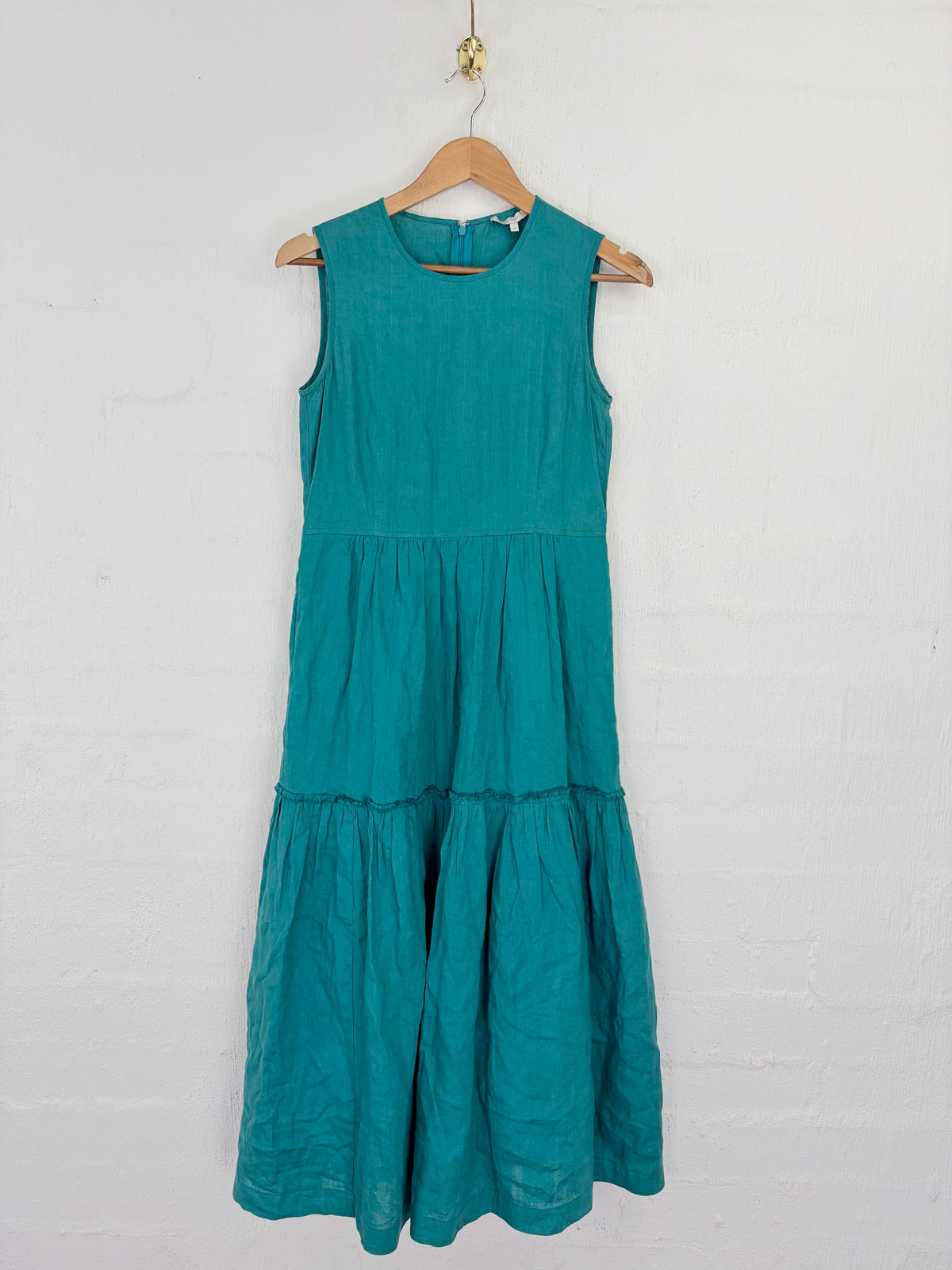 A sleeveless tiered maxi dress in teal green hangs on a wooden hanger against a white painted brick wall.