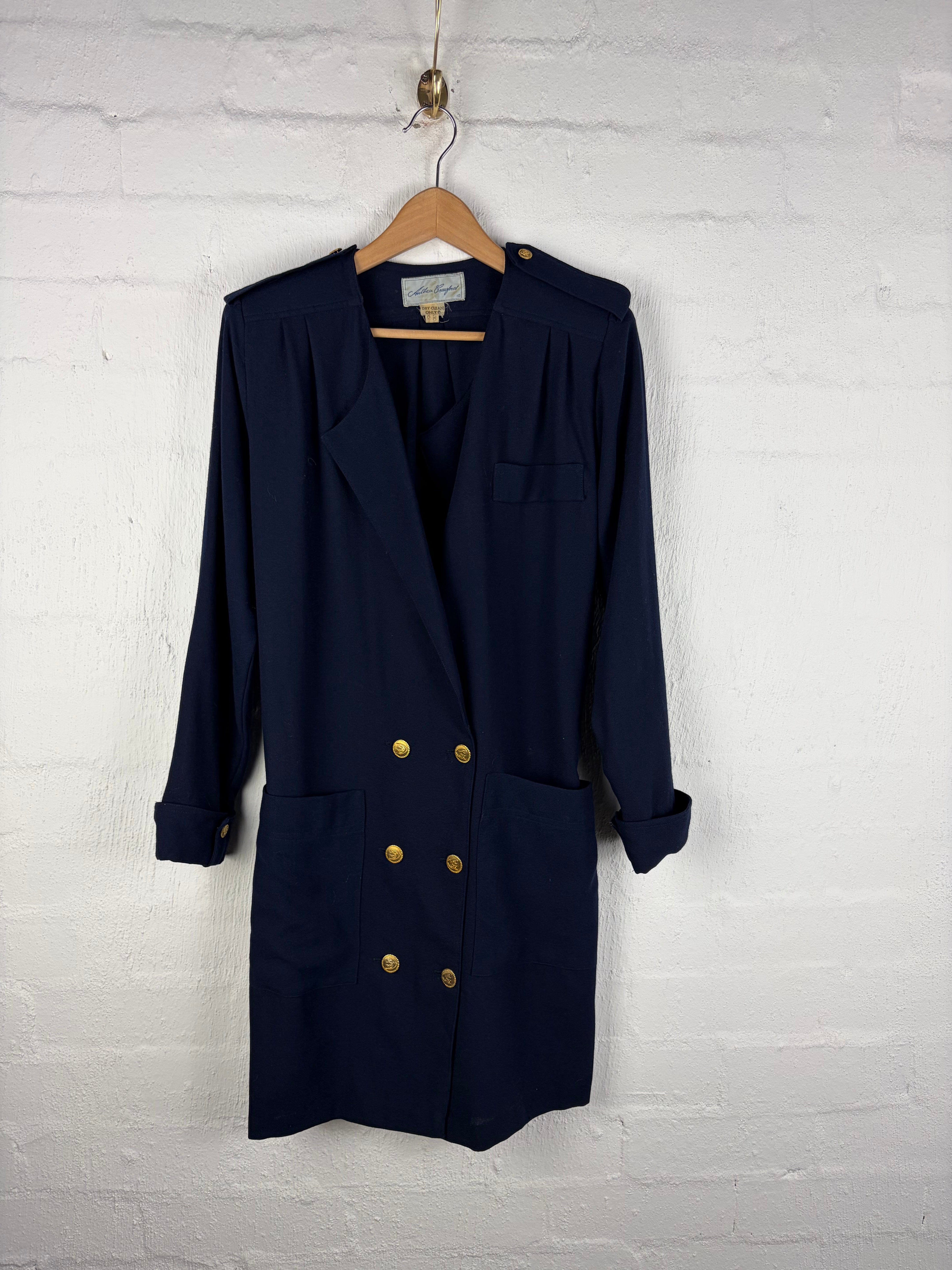 A navy blue double-breasted blazer with gold buttons, hanging on a wooden hanger against a white painted brick wall.