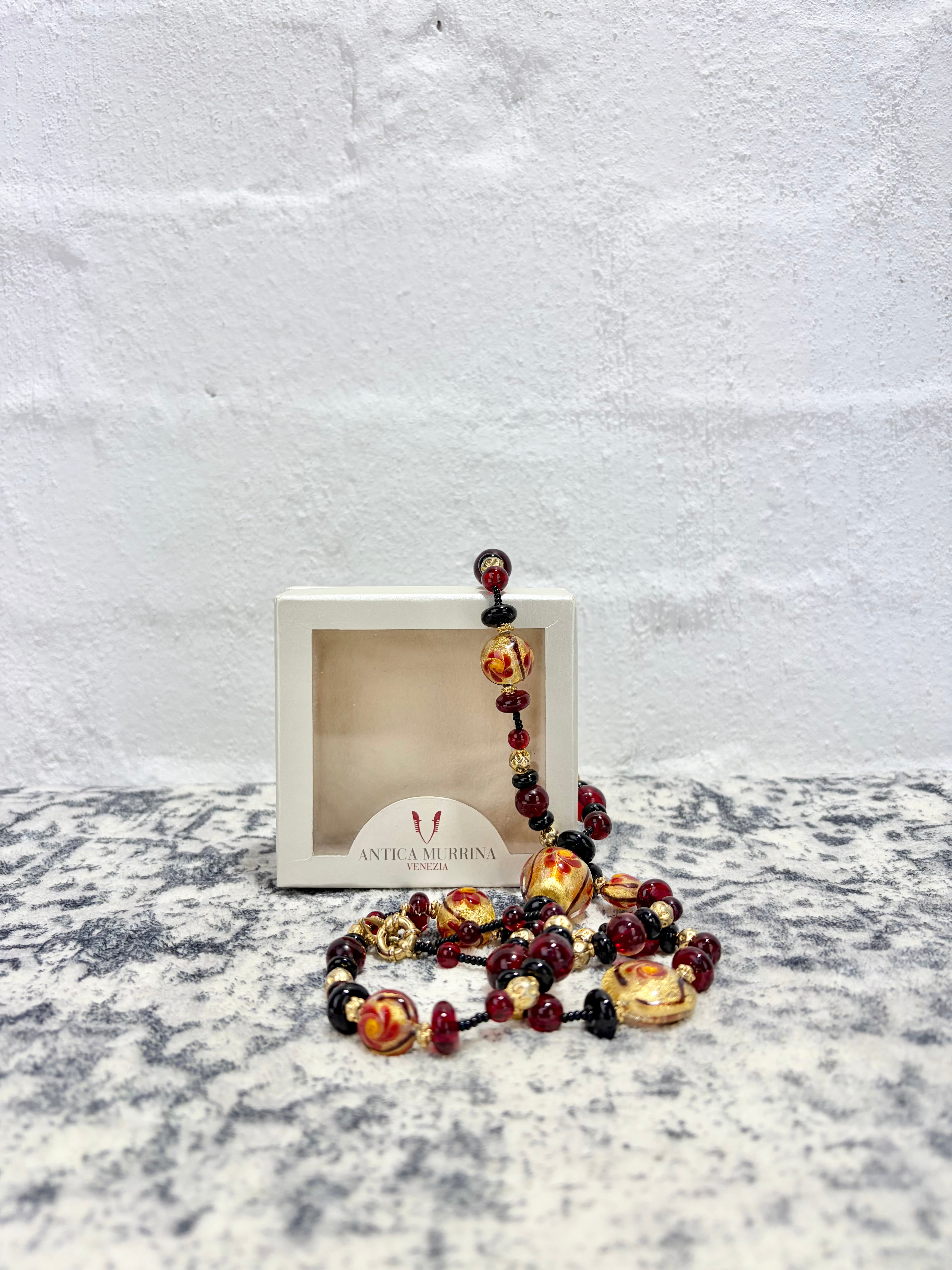 A beaded necklace with large gold-toned and amber-red beads, displayed in front of a small white jewelry box on a mottled gray surface against a white brick wall.
