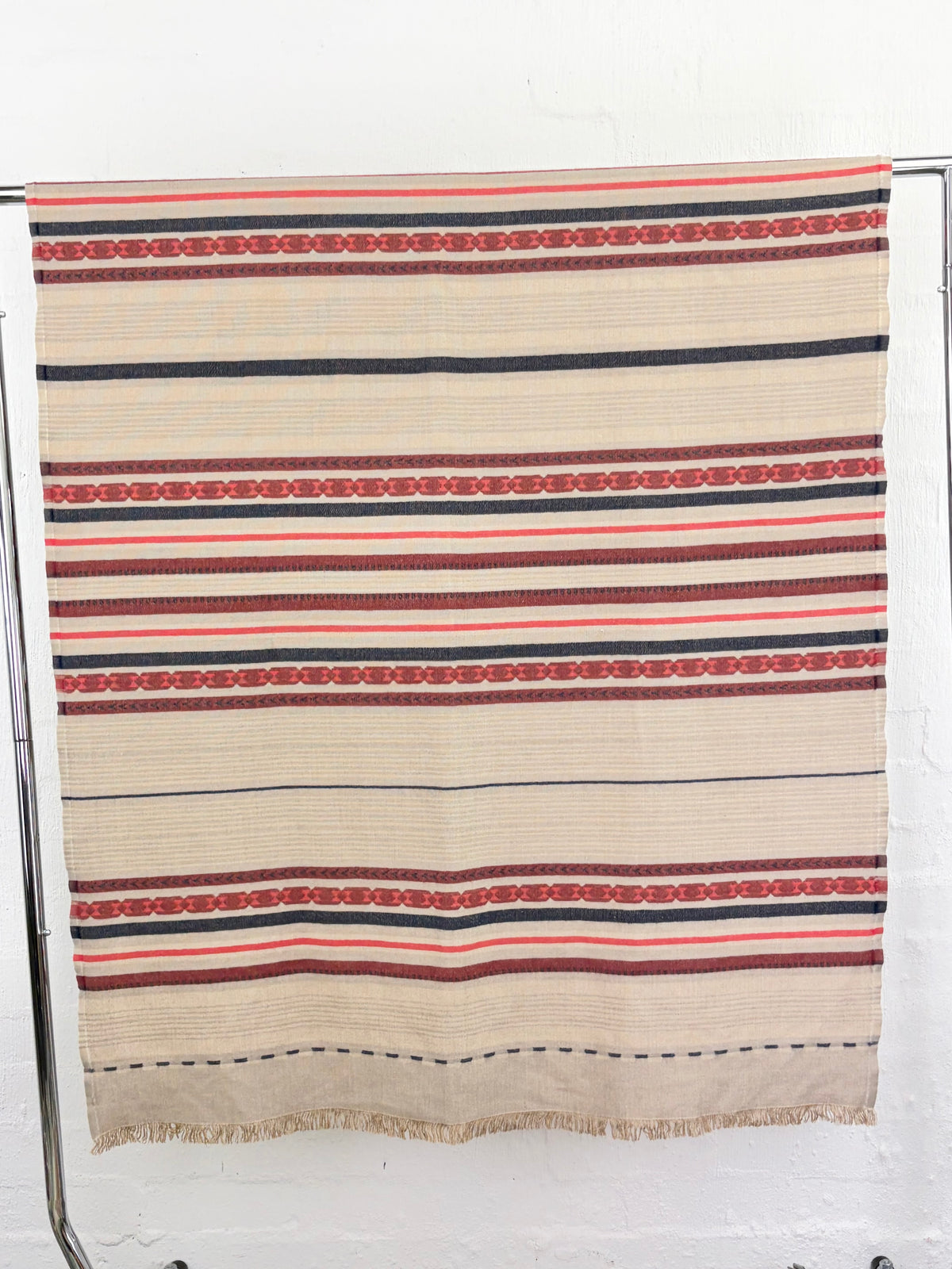 A woven textile blanket or throw with horizontal stripes in beige, red, black, and orange, hanging on a metal rack against a white wall.