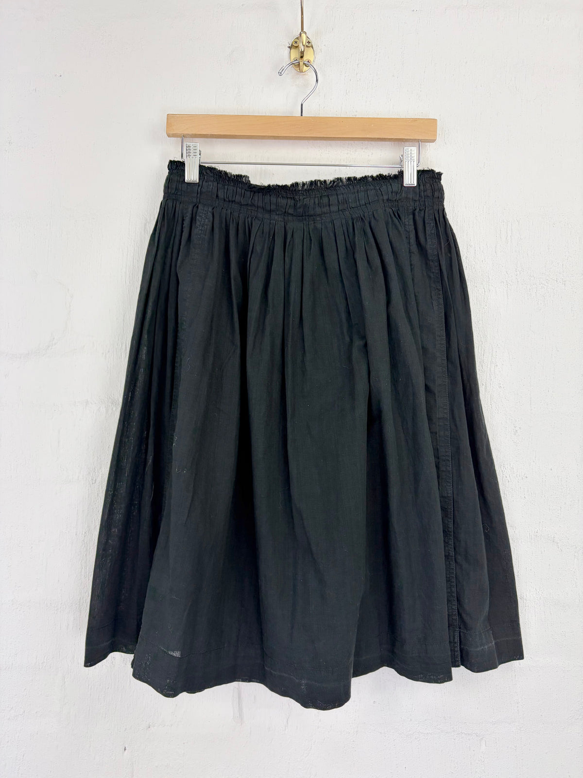 A black pleated linen skirt hanging from a wooden hanger against a white textured wall.