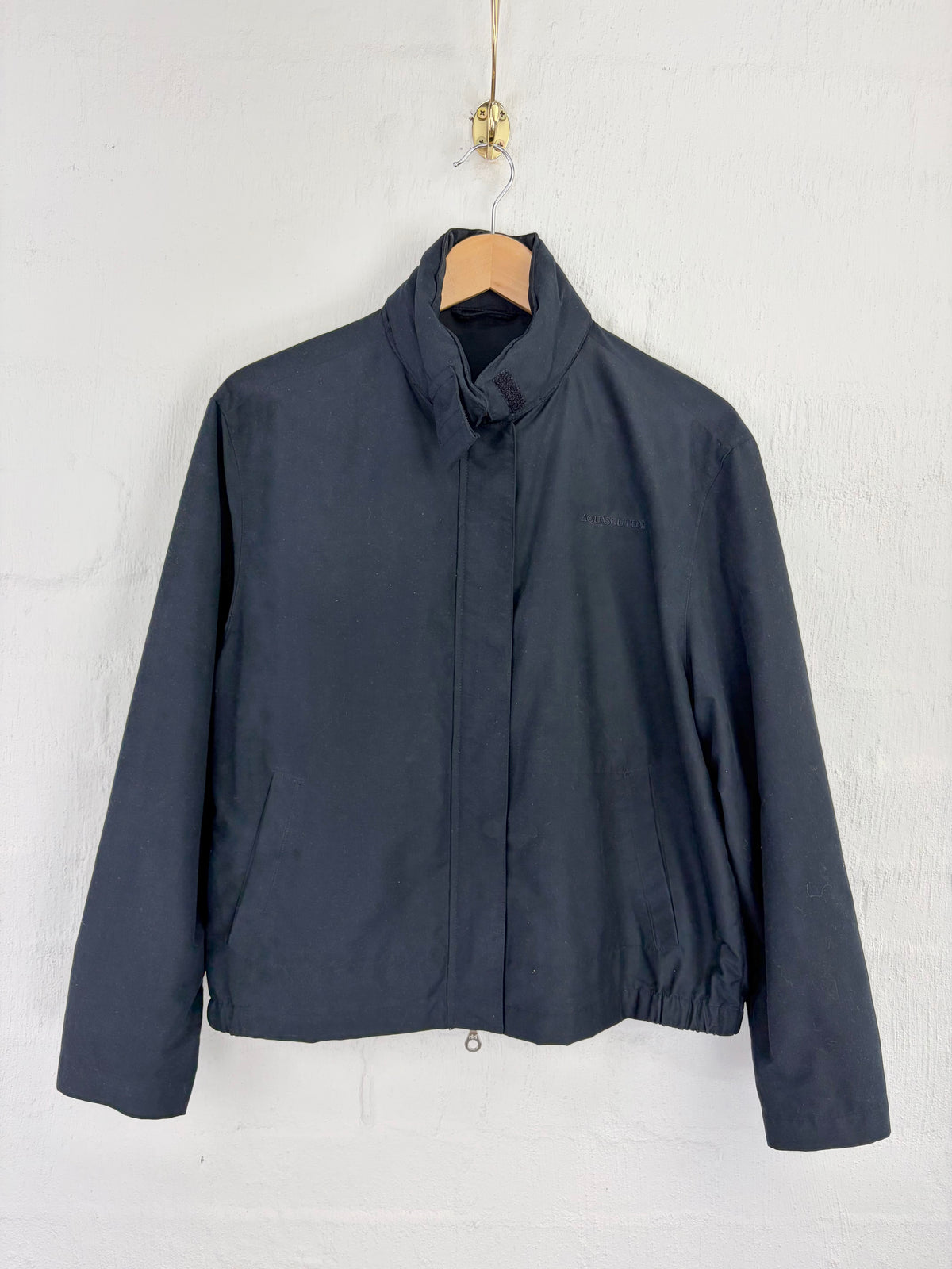 A dark navy blue lightweight jacket with a high stand-up collar, hanging on a wooden hanger against a white textured wall.