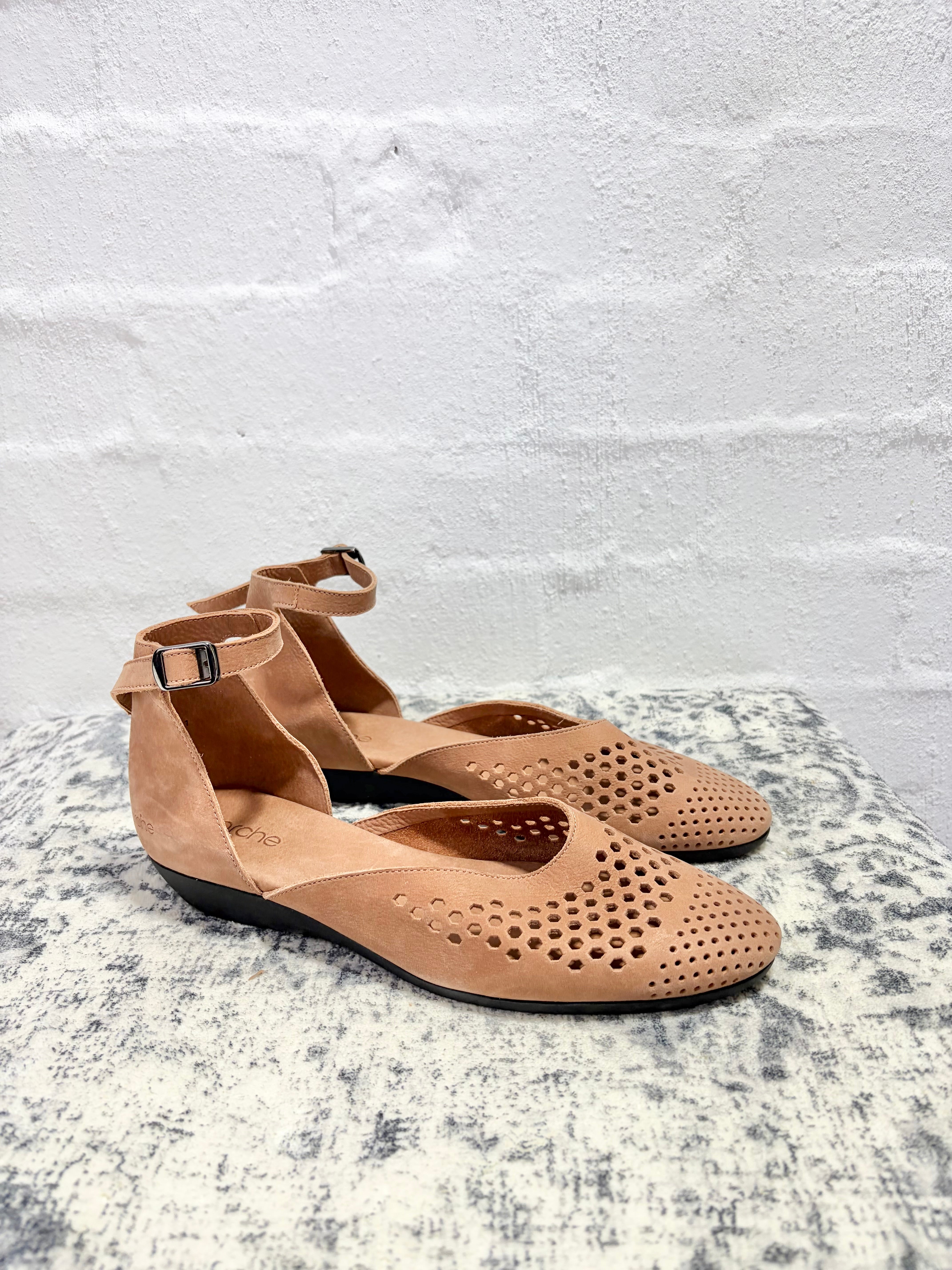 A pair of tan perforated flat sandals with ankle straps, displayed on a textured gray stone surface against a white brick wall.
