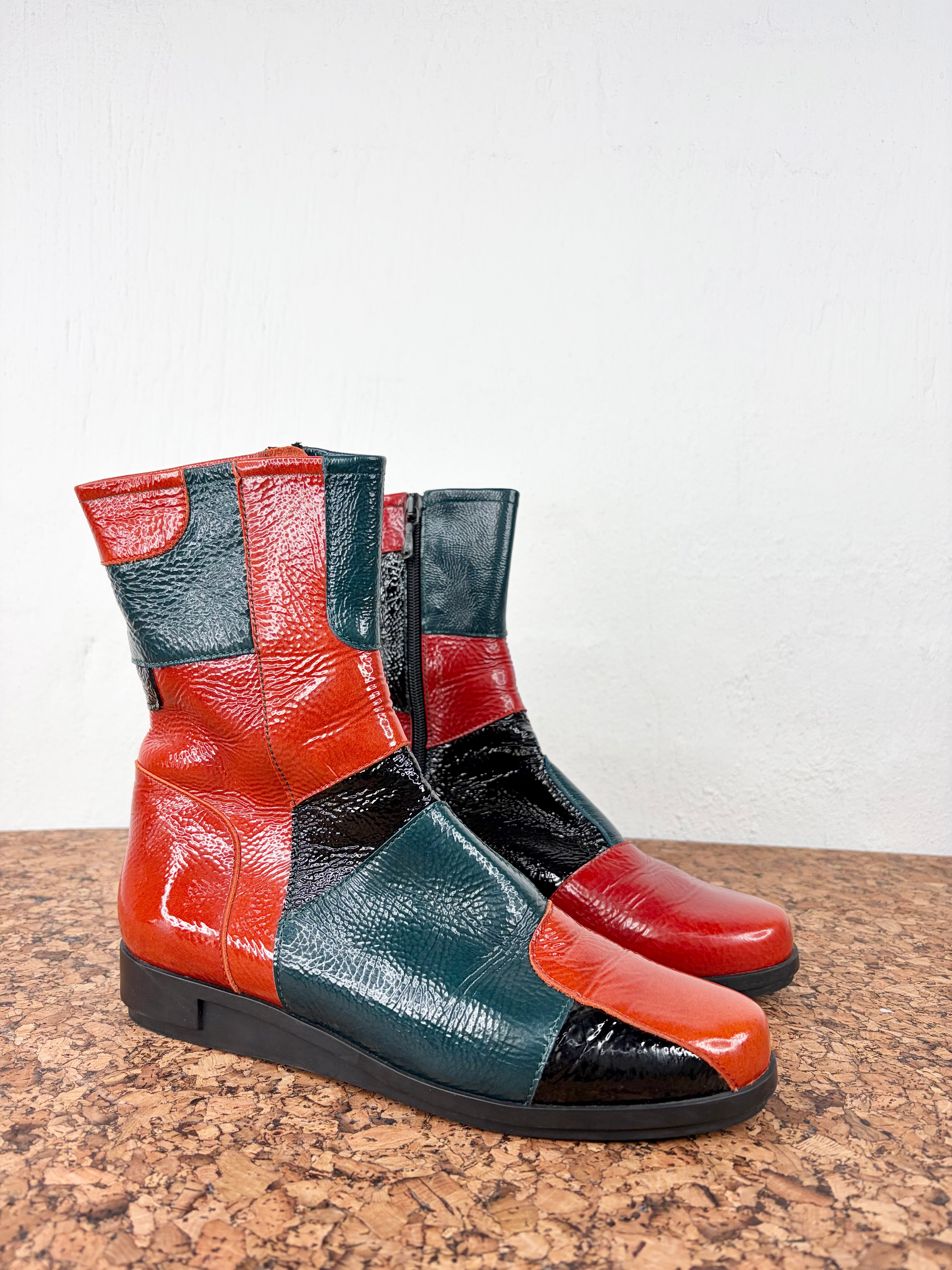 A pair of mid-calf patchwork leather boots with a glossy finish, featuring color-blocked panels of red, dark teal, and black, resting on a cork surface against a white wall.