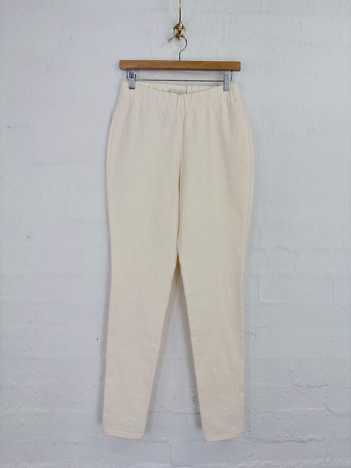 Cream-colored cropped pants hanging from a wooden hanger against a white painted brick wall.