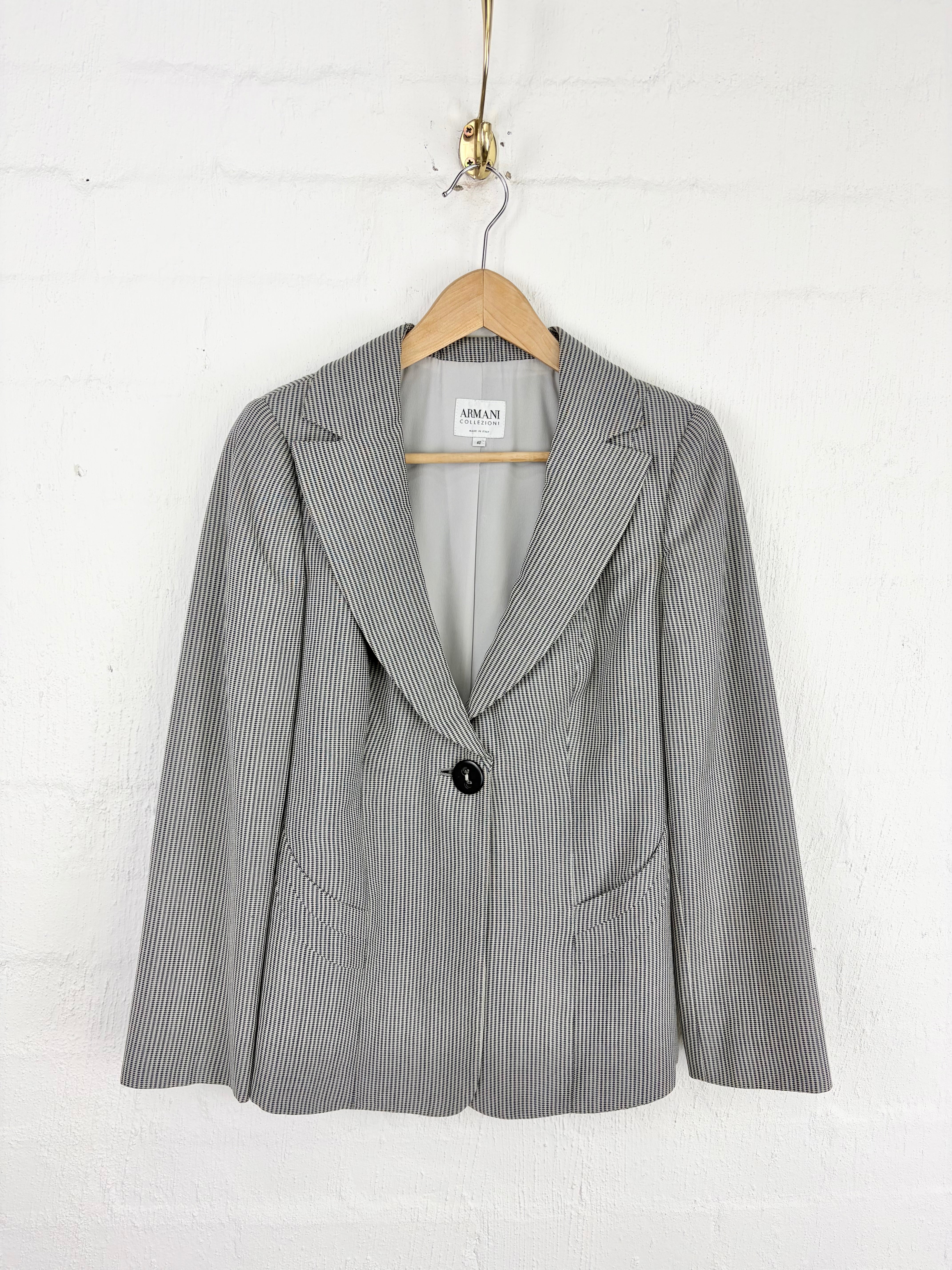 A gray pinstriped blazer with a single black button, hanging on a wooden hanger against a white brick wall.