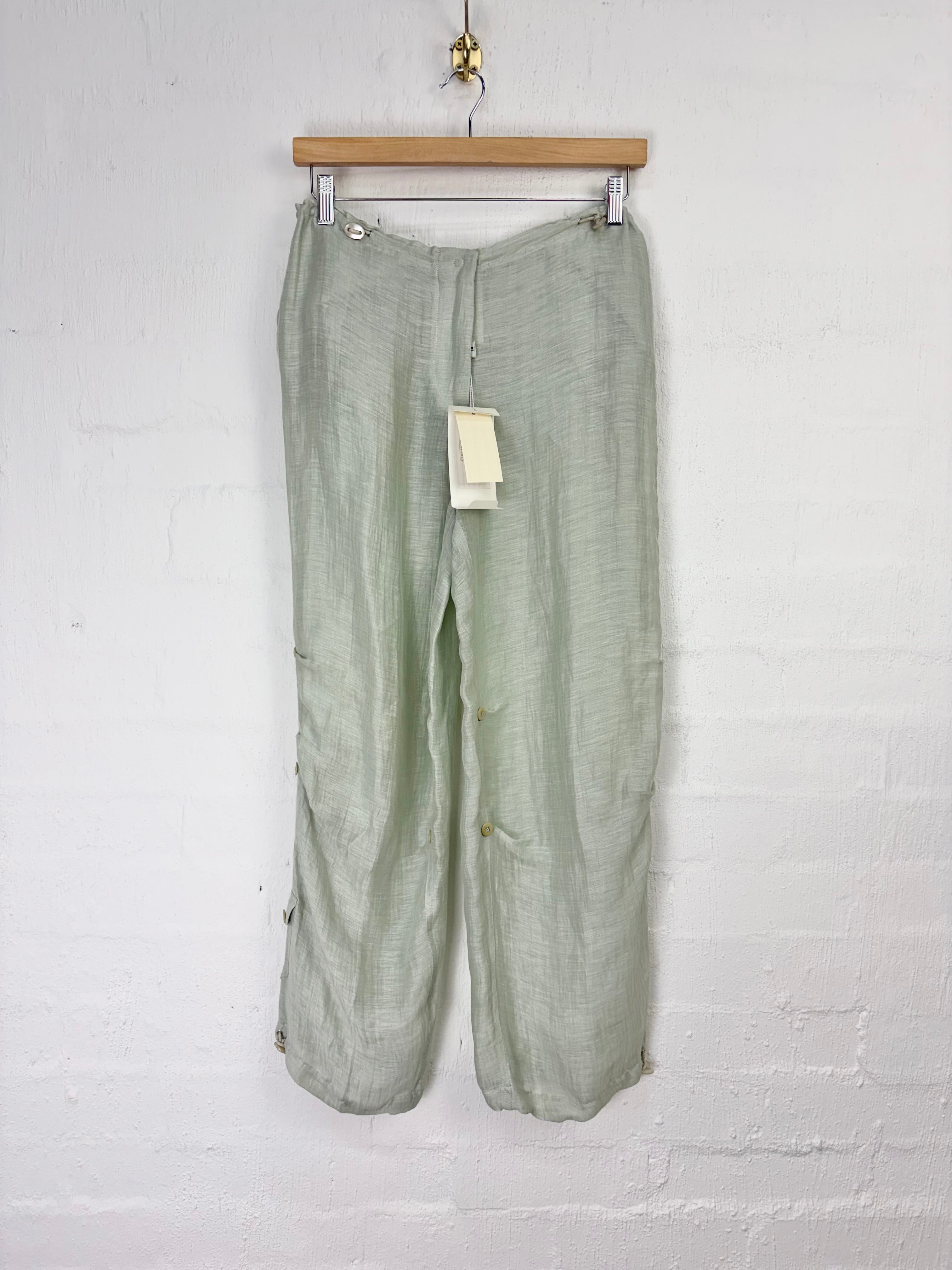 Light sage green linen cropped pants hanging from a wooden hanger against a white painted brick wall.