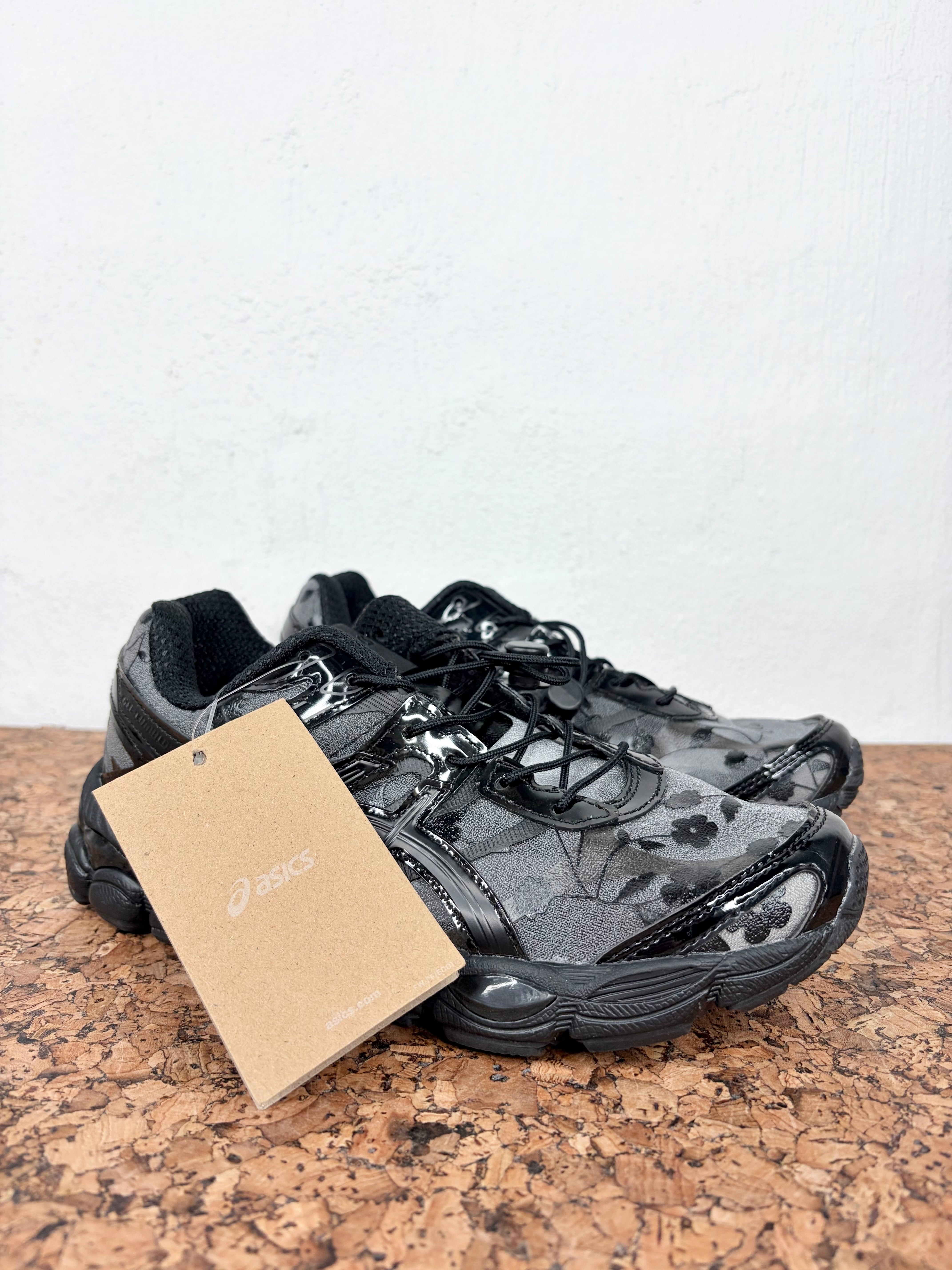 A pair of Asics athletic sneakers in black and gray with a glossy finish, resting on a cork surface against a white wall. A brown paper tag with the Asics logo is attached to the front shoe.