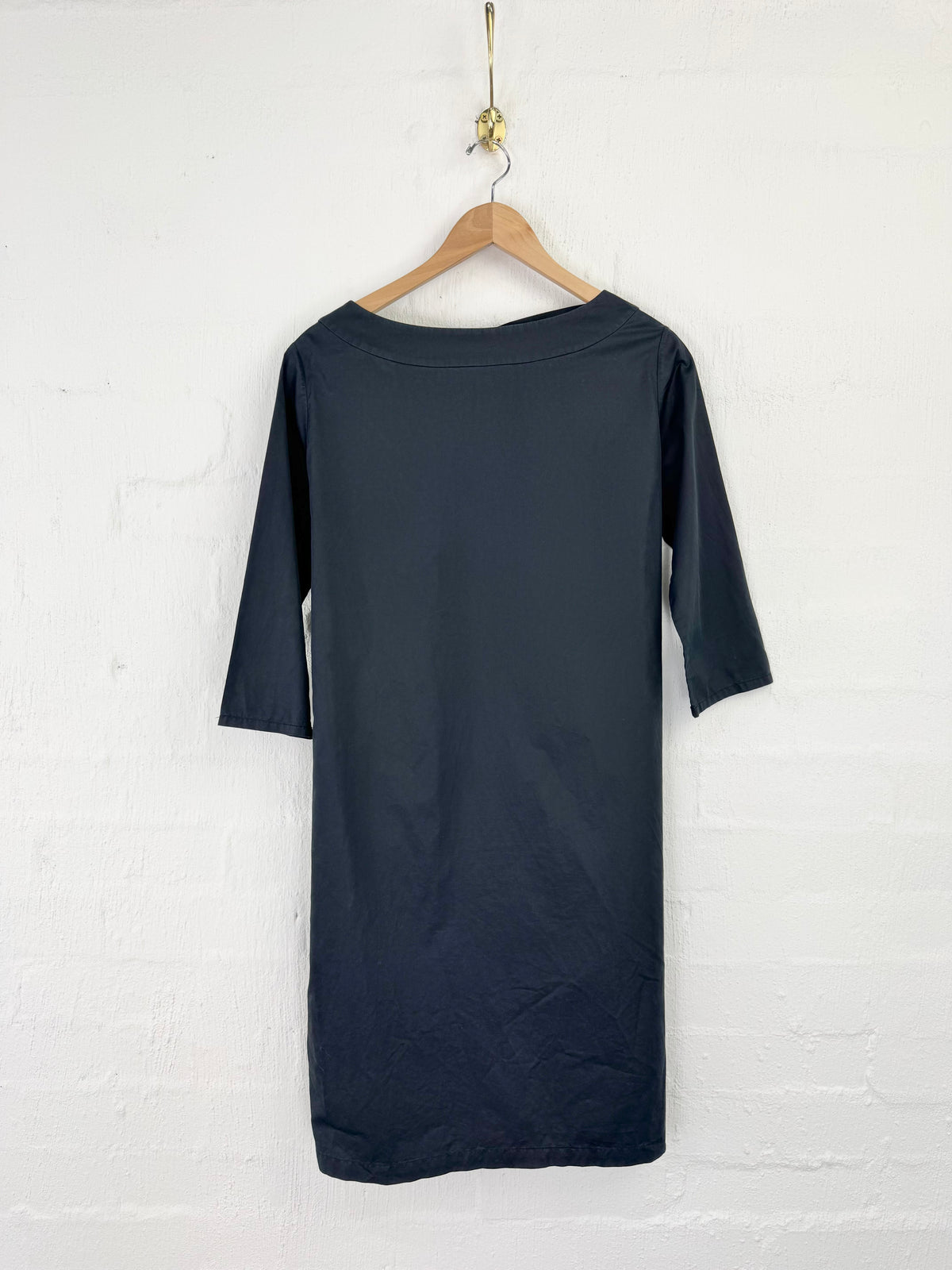 A dark navy blue dress with three-quarter sleeves hanging on a wooden hanger against a white brick wall.