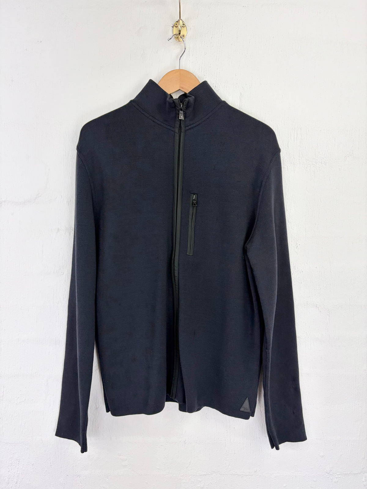 A dark navy long-sleeve quarter-zip pullover jacket hanging on a wooden hanger against a white painted brick wall.