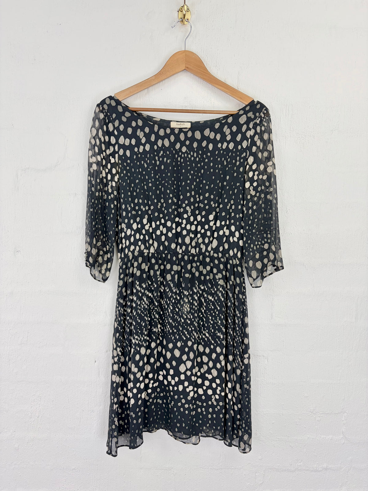 A sheer black midi dress with an abstract white spotted pattern, hanging on a wooden hanger against a white painted brick wall.
