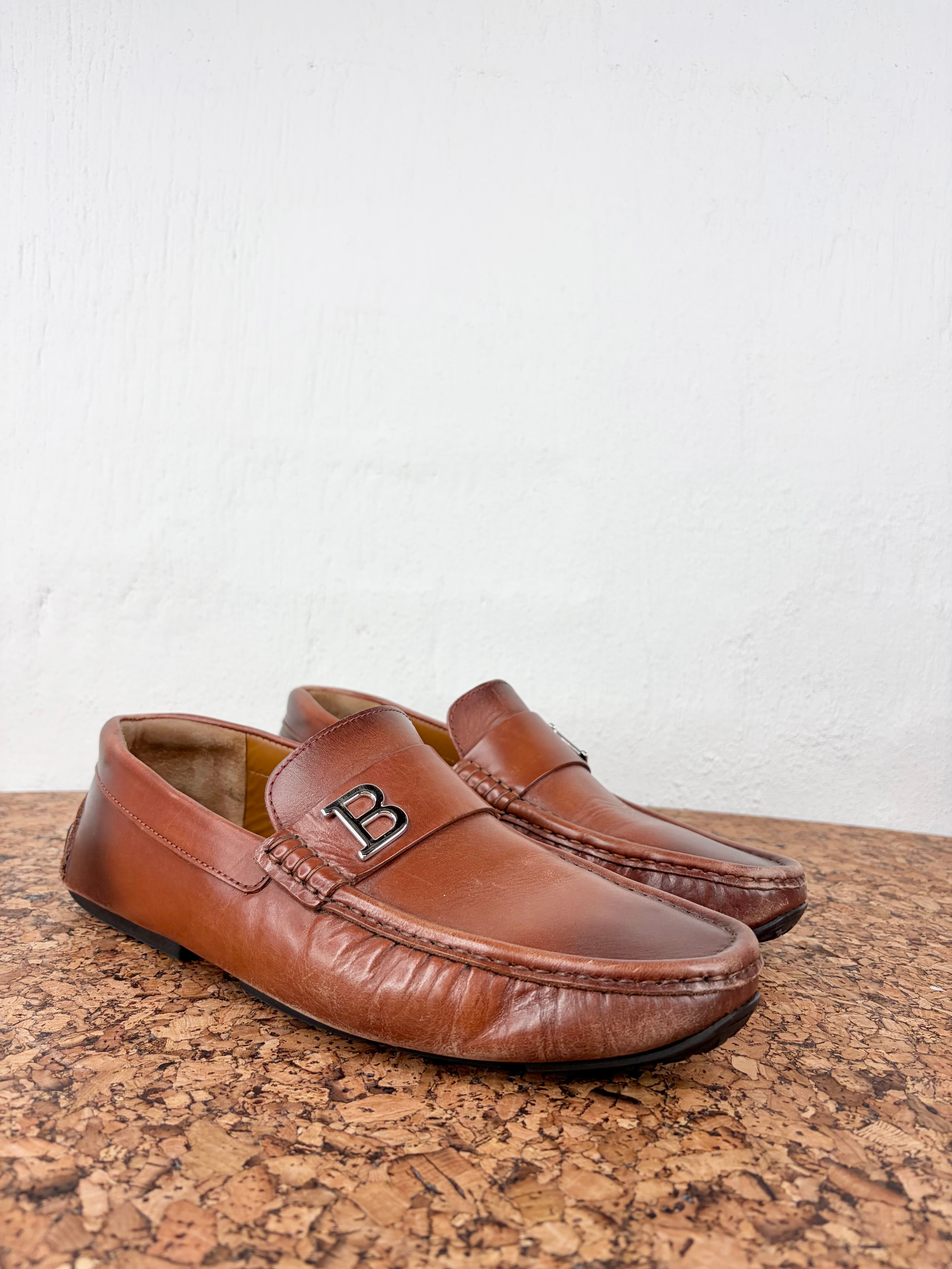 A pair of brown leather penny loafers with a prominent black 'B' logo on the vamp, displayed on a cork surface against a white wall.