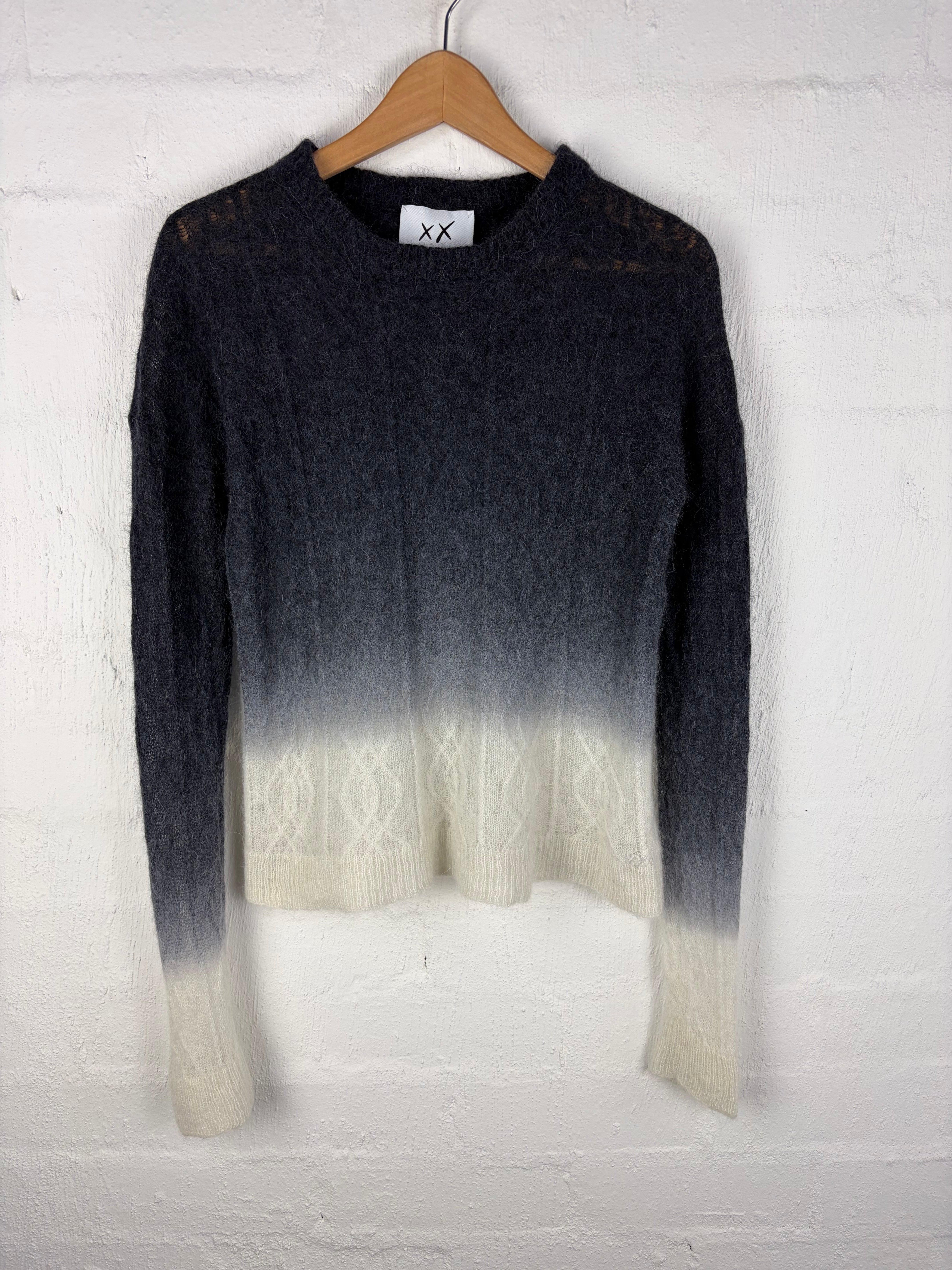 A long-sleeve knit sweater with a dark charcoal-to-cream ombré gradient, hanging on a wooden hanger against a white painted brick wall.