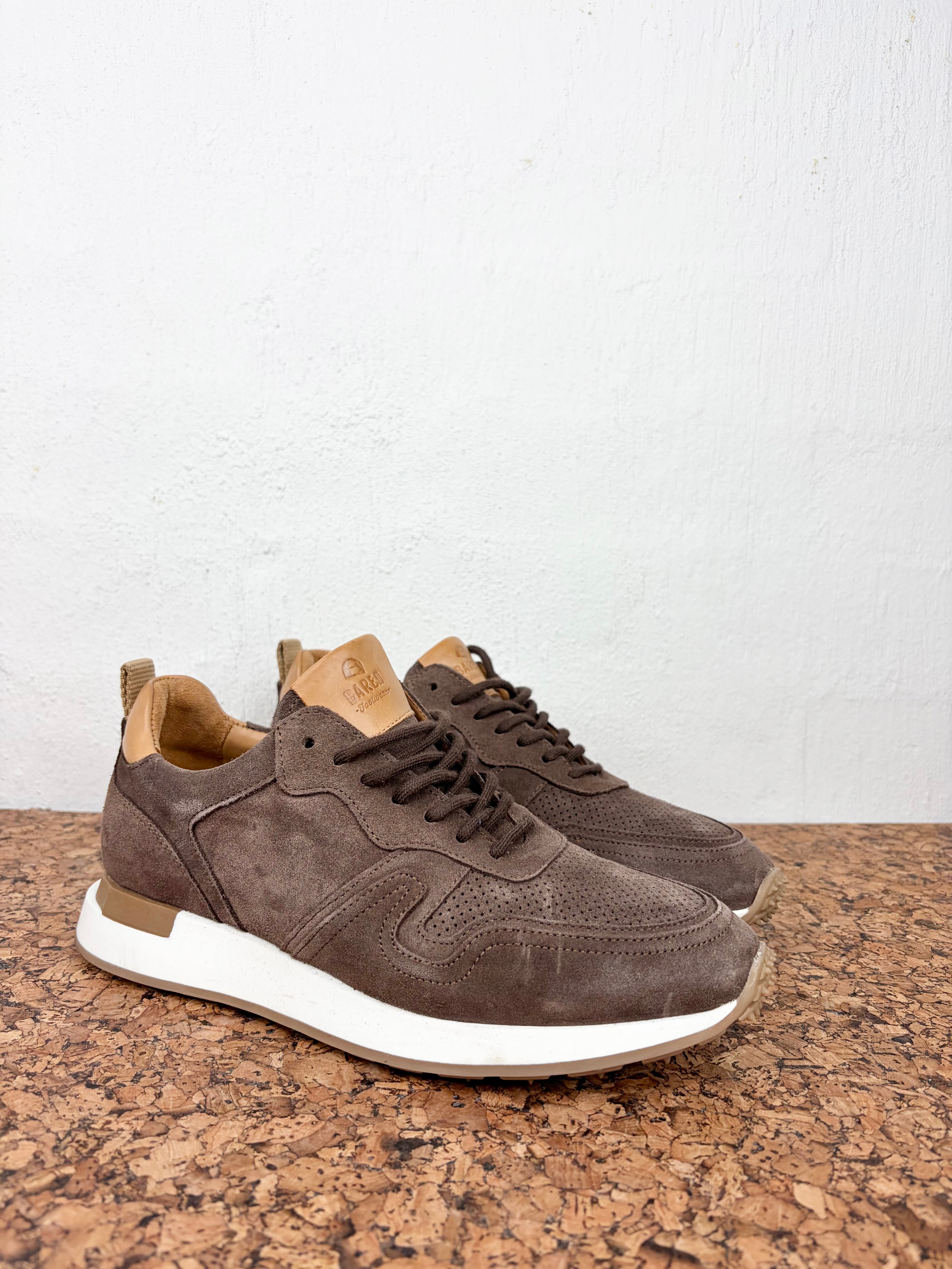 A pair of brown suede and perforated leather sneakers with white midsoles, displayed on a textured cork surface against a white wall.