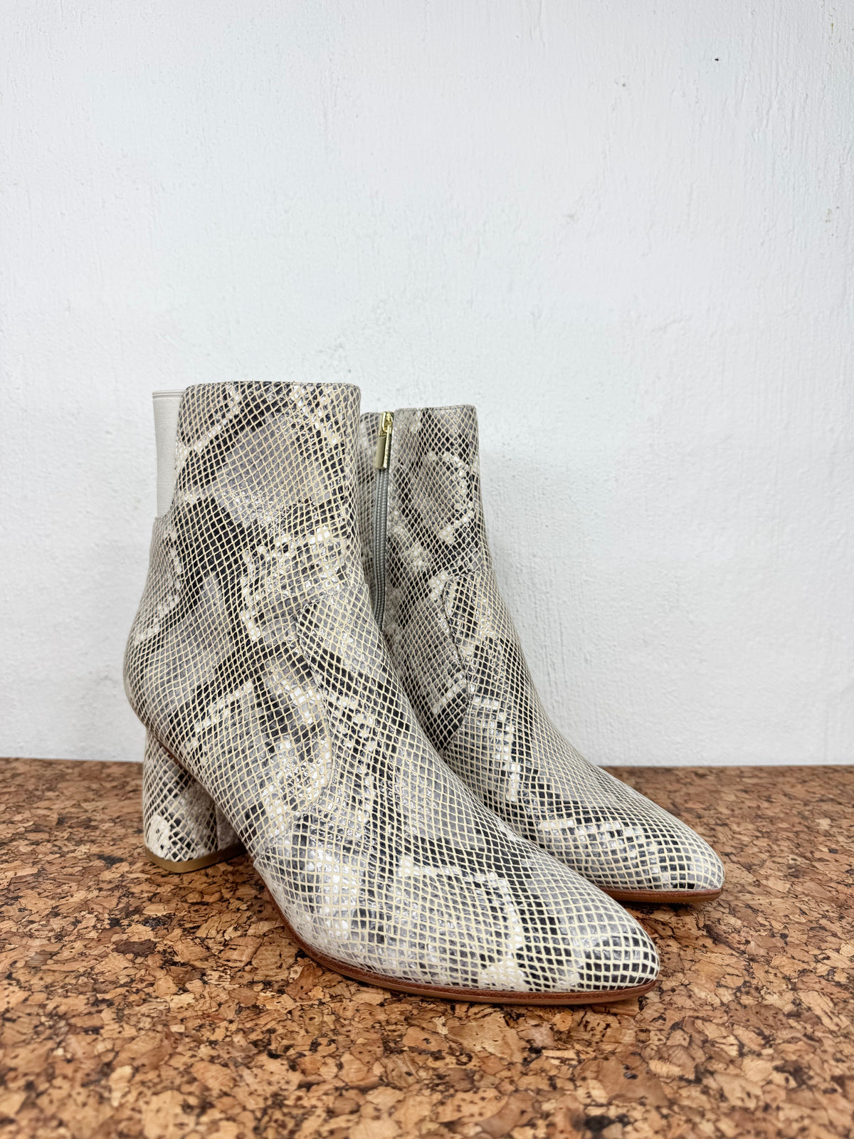 A pair of snakeskin-patterned ankle boots with a pointed toe and block heel, displayed on a cork surface against a white wall.