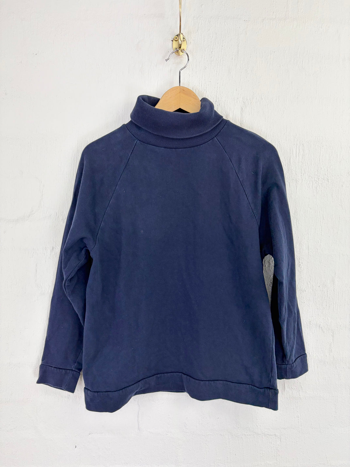 A navy blue long-sleeve turtleneck top hanging on a wooden hanger against a white textured wall.