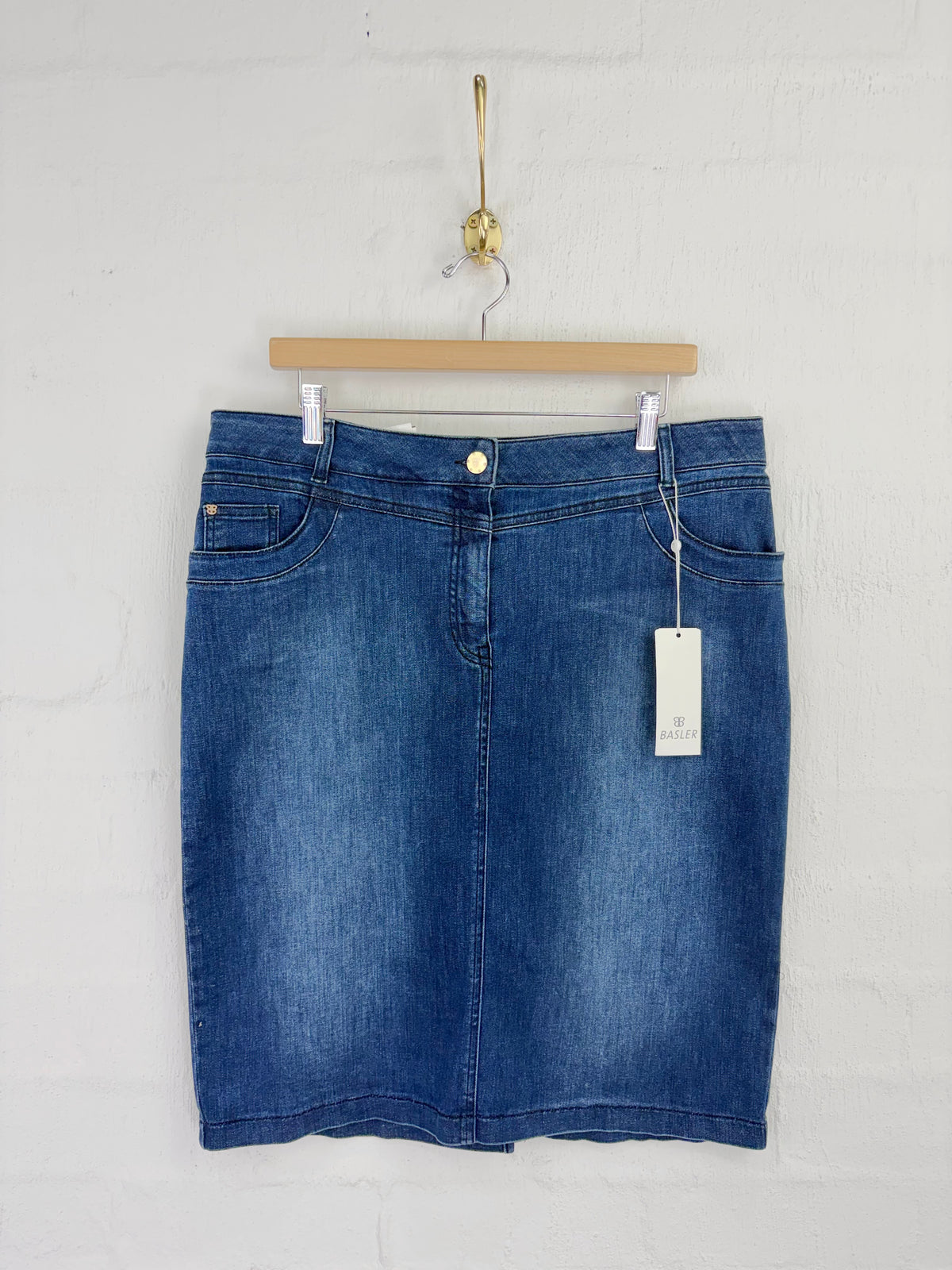 A blue denim mini skirt hanging from a wooden hanger against a white painted brick wall. The skirt features a classic five-pocket design, a button closure, and a white hangtag attached to the side.