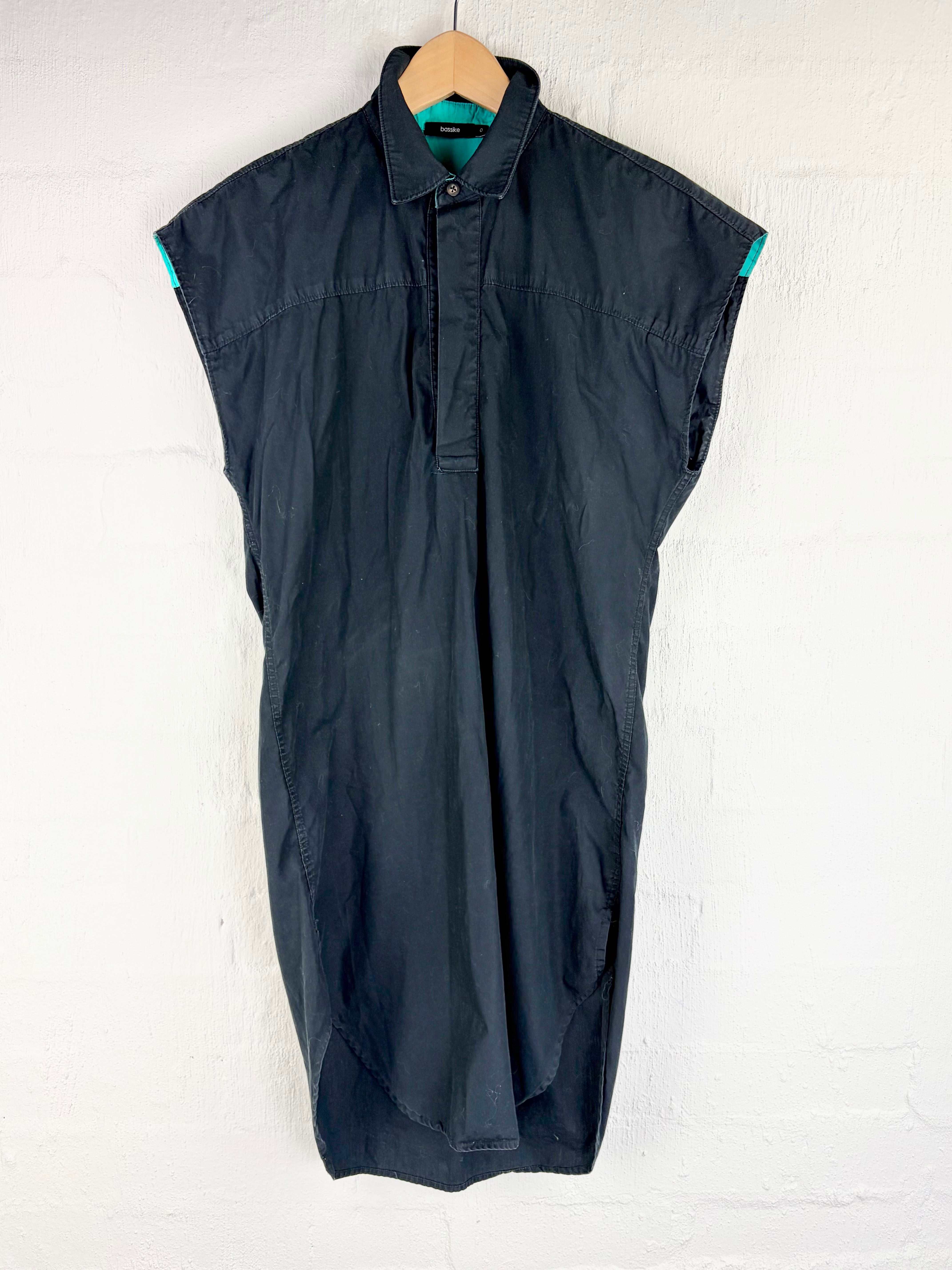 A sleeveless black button-up shirt dress with a pointed collar, hanging on a wooden hanger against a white painted brick wall. Small teal accents are visible at the inner collar and sleeve cuffs.