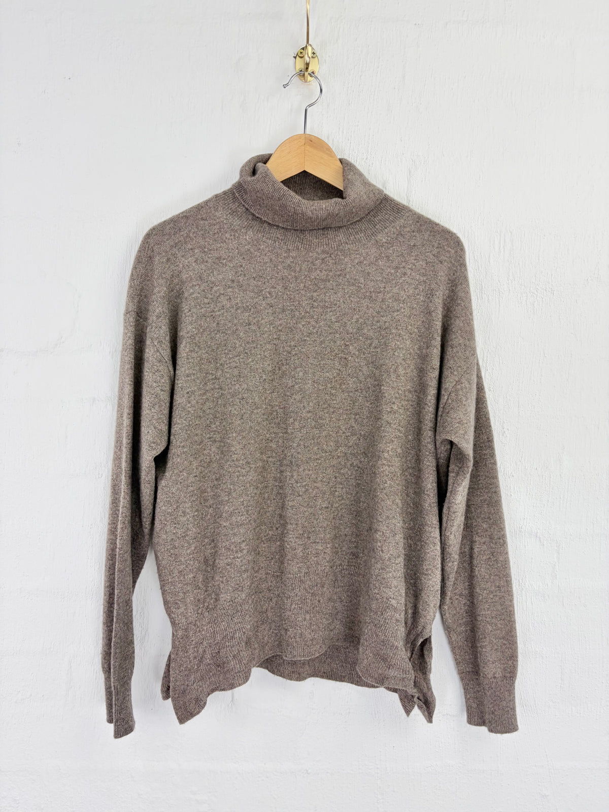 A taupe brown knit turtleneck sweater hanging on a wooden hanger against a white painted brick wall.