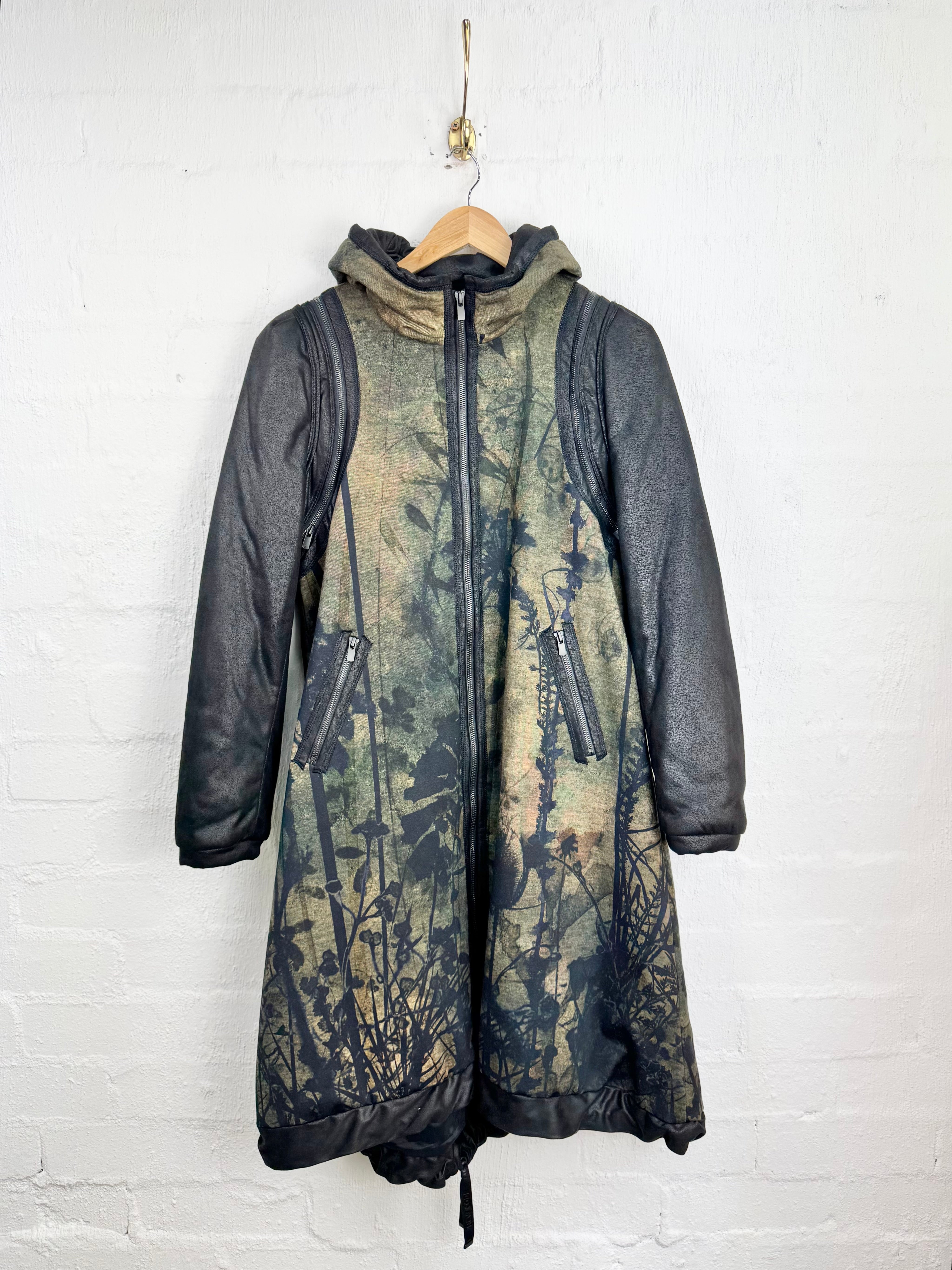 A hooded mid-length jacket hanging on a wooden hanger against a white painted brick wall. The jacket has dark charcoal gray quilted sleeves and hood trim, with a central front zipper and a body panel featuring an abstract, distressed tree-like print in muted olive, beige, and dark gray tones.