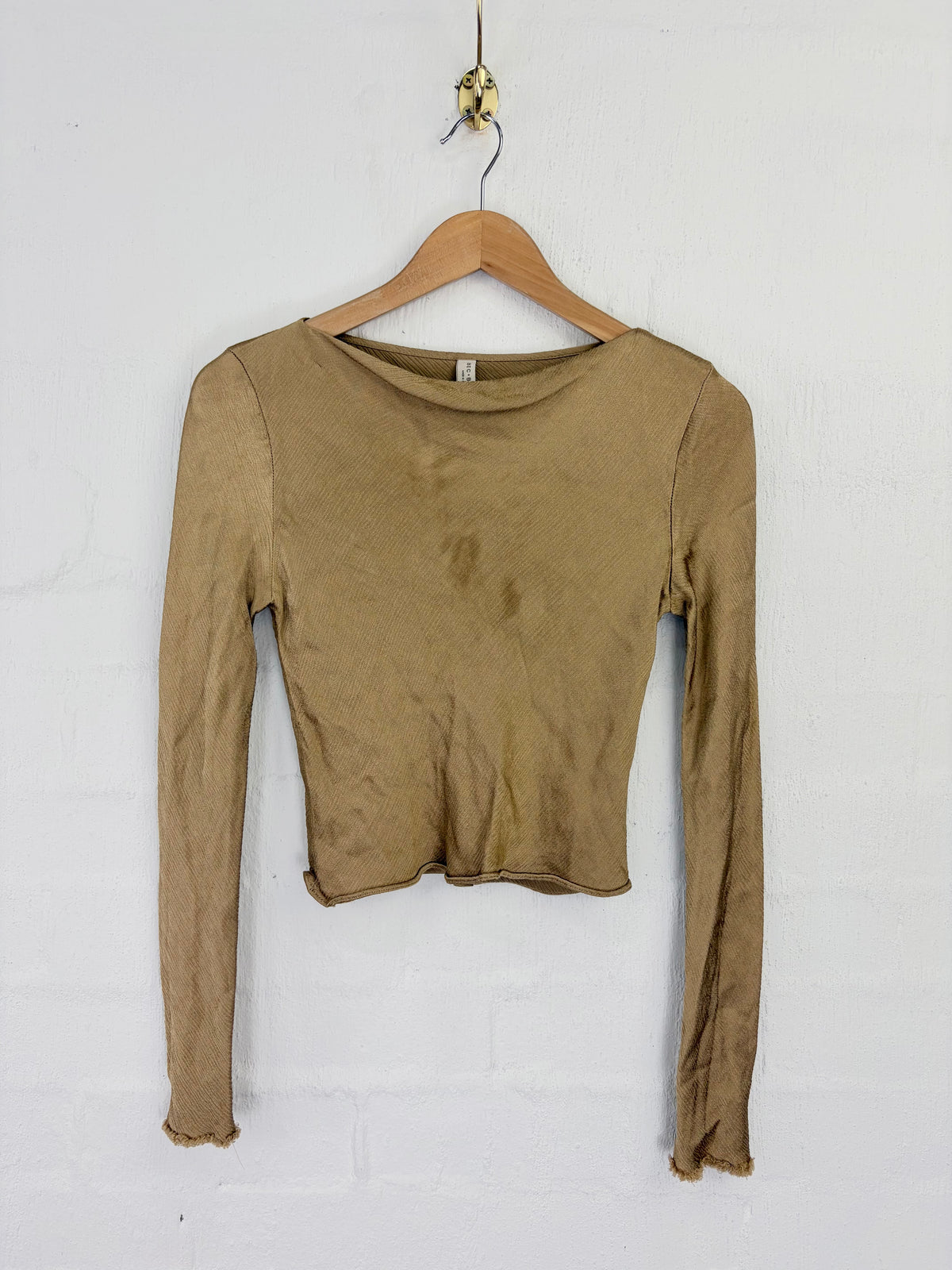 A long-sleeve cropped knit top in a muted khaki-brown color, hanging on a wooden hanger against a white painted brick wall.