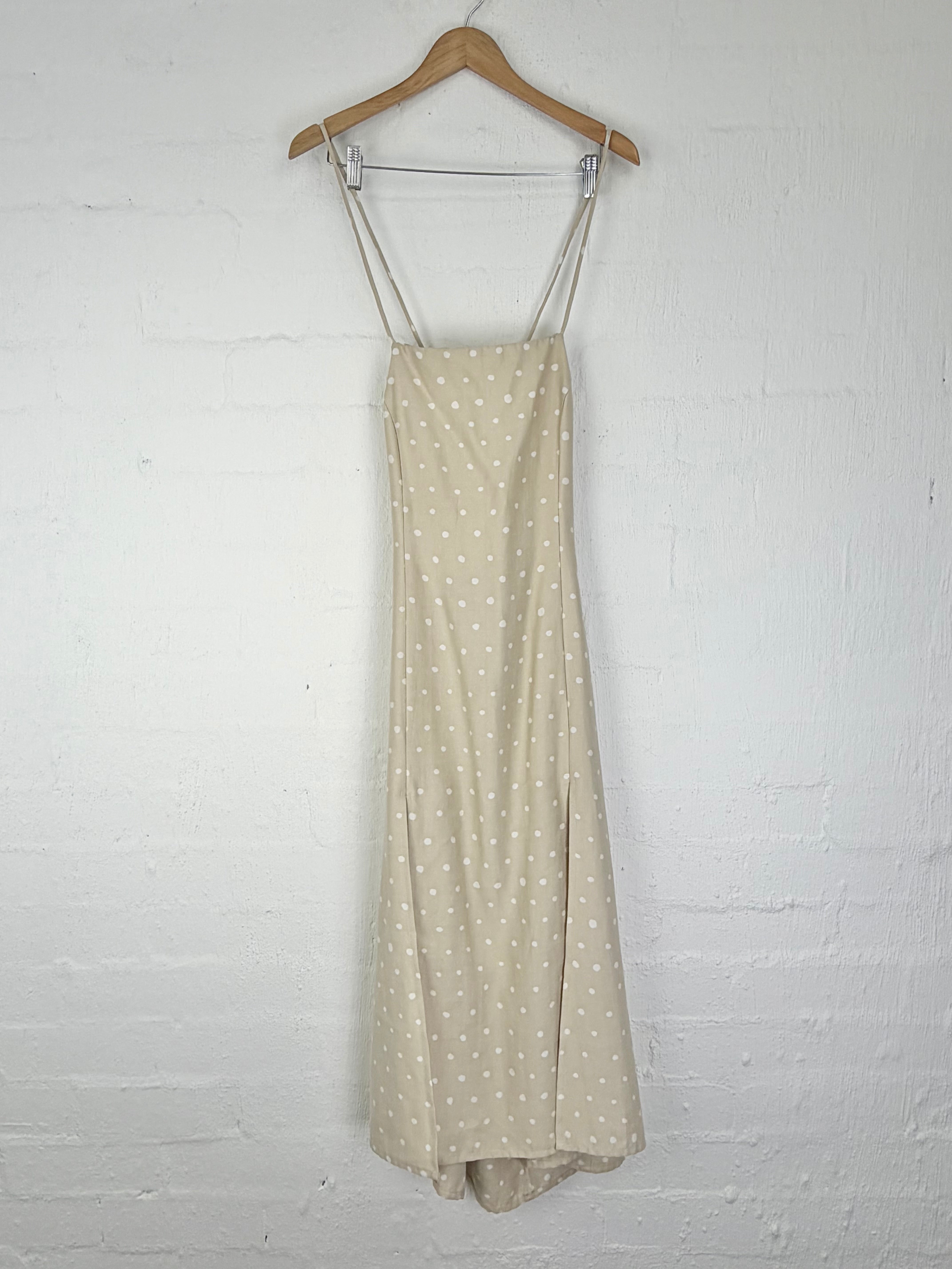 A light beige maxi dress with white polka dots hanging from a wooden hanger against a white painted brick wall.