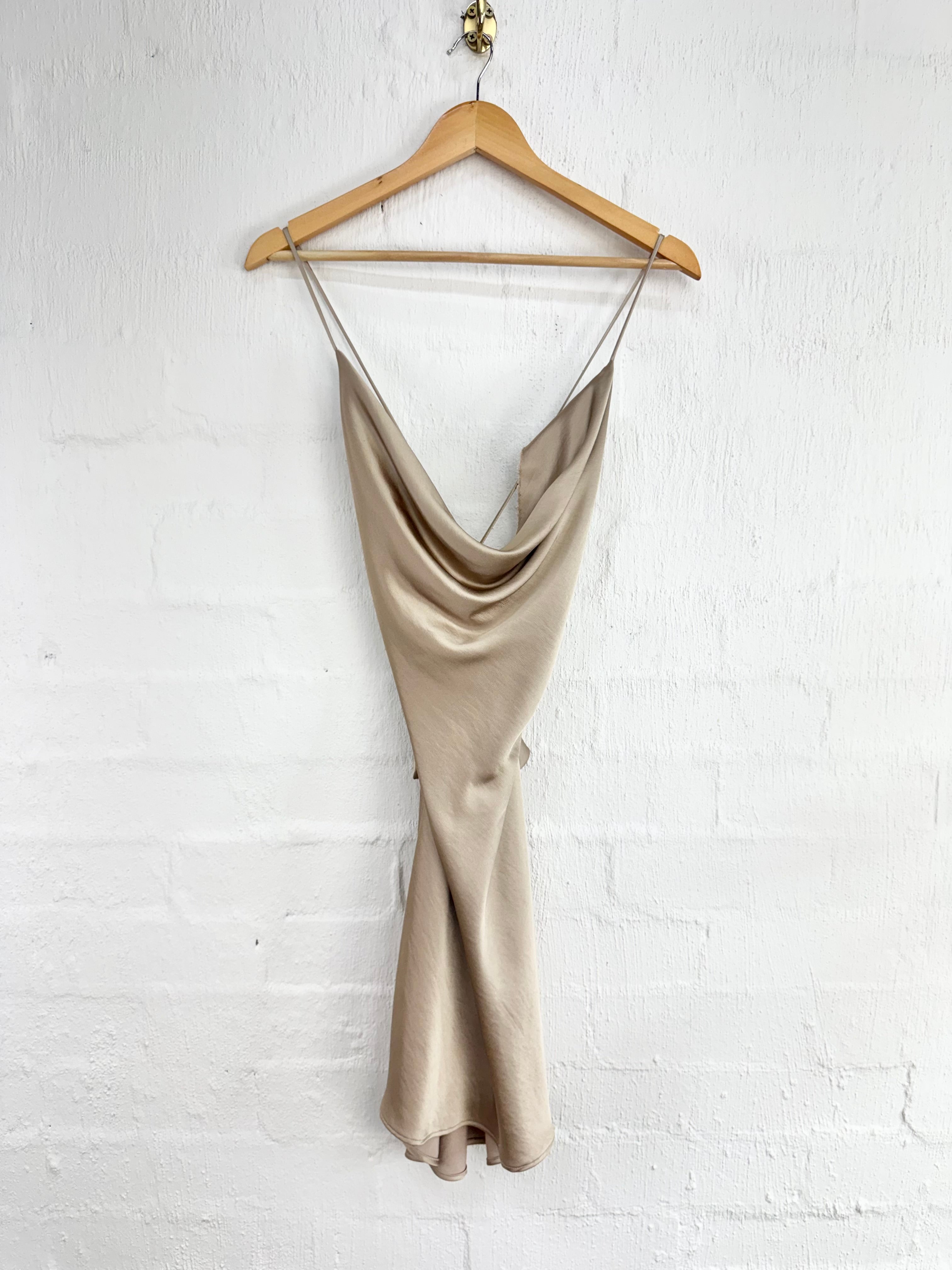 A beige satin slip dress with a cowl neckline and thin spaghetti straps, hanging on a wooden hanger against a white painted brick wall.