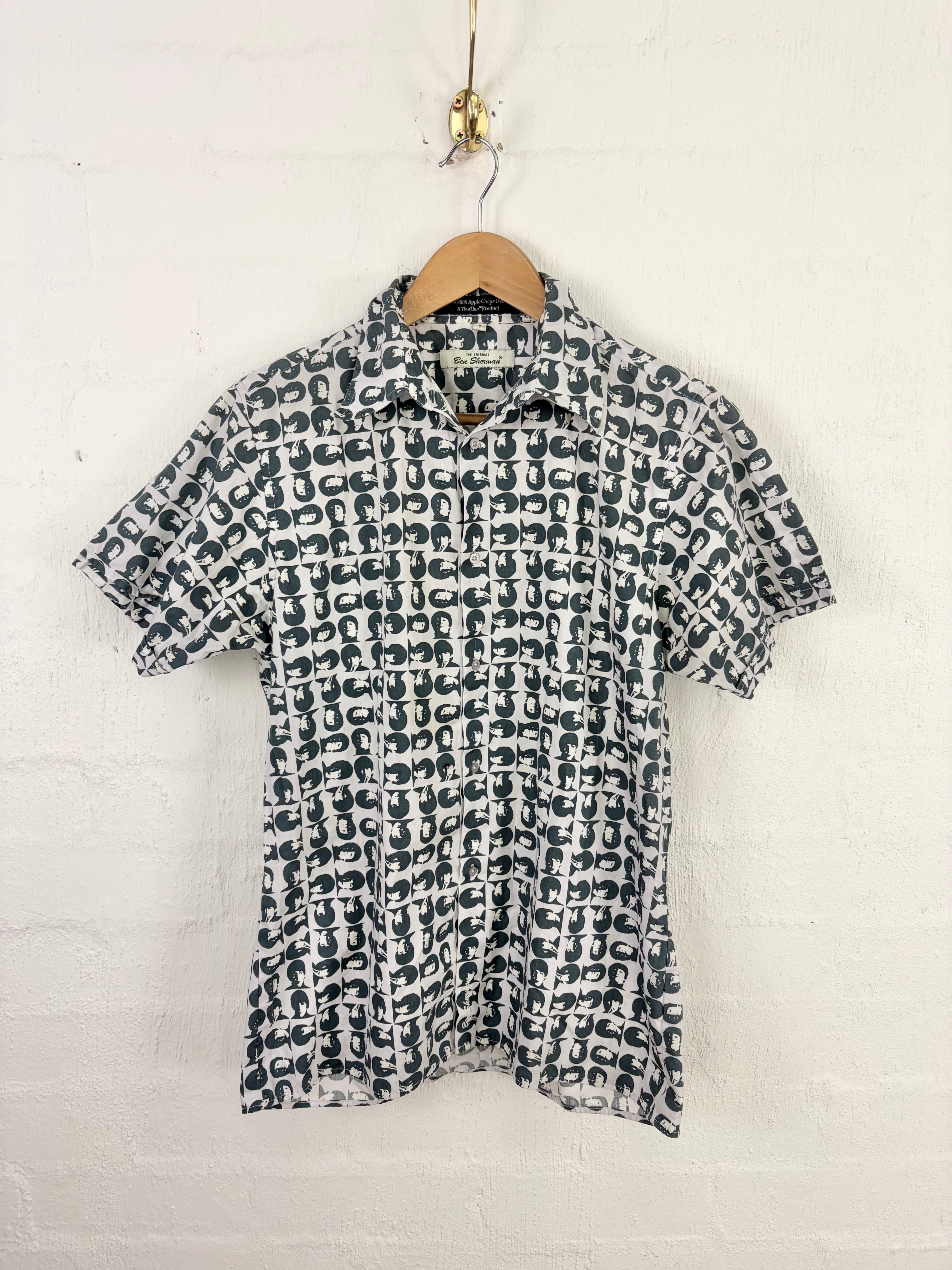 A short-sleeve button-up shirt with an all-over abstract black and off-white pattern, hanging on a wooden hanger against a white painted brick wall.