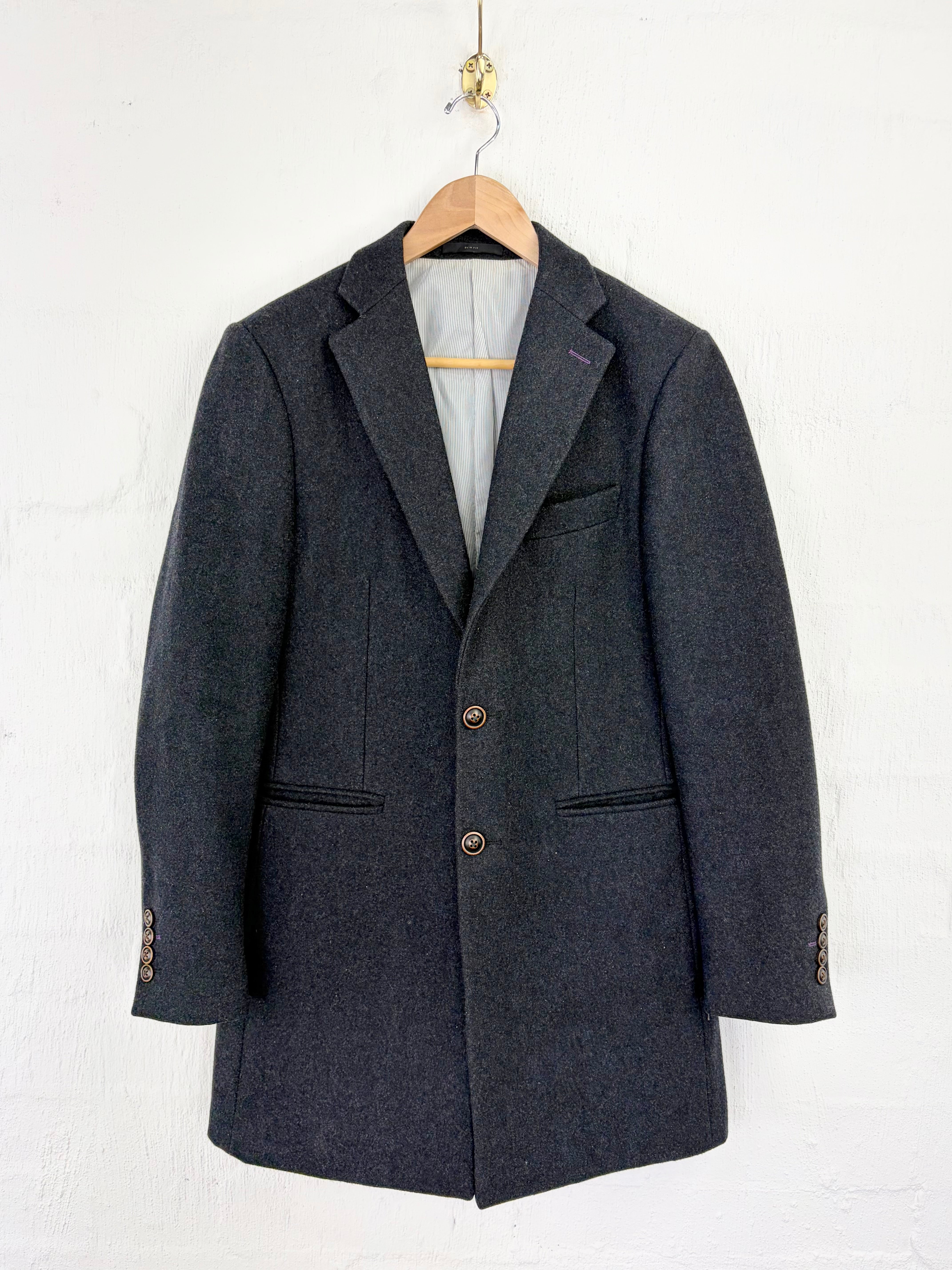 A dark gray textured wool blazer hanging on a wooden hanger against a white painted brick wall.