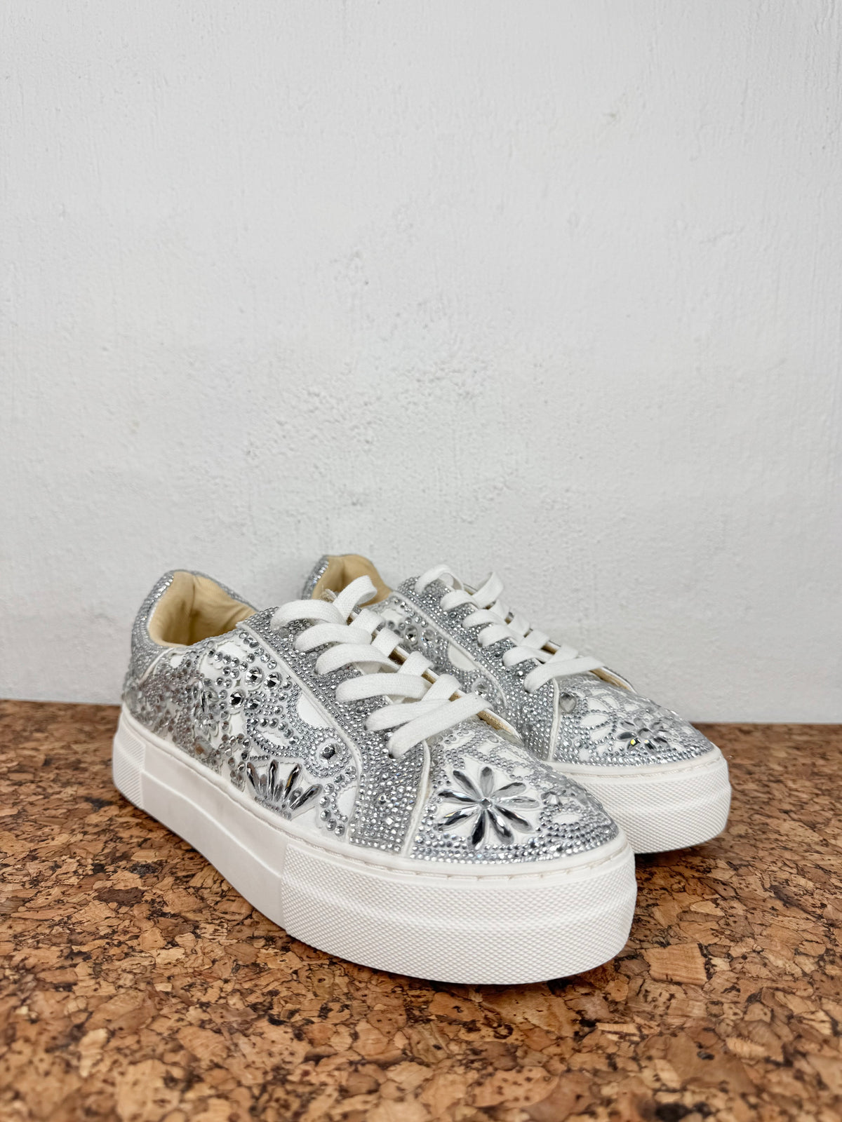 A pair of low-top platform sneakers covered in silver rhinestones with white floral embroidery, white laces, and chunky white soles, displayed on a cork surface against a white wall.