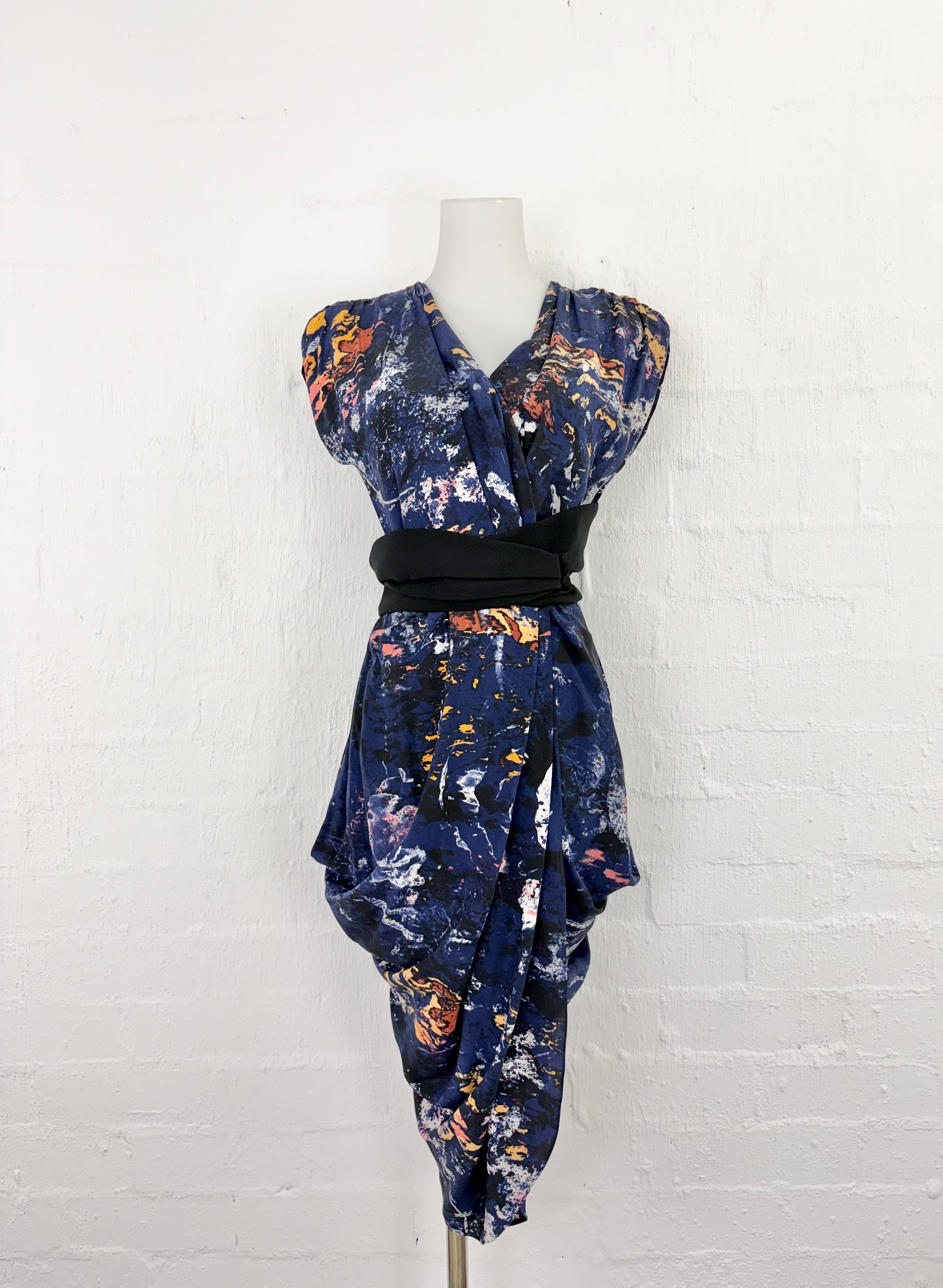 A navy blue wrap dress with a colorful abstract floral print, styled on a mannequin with a black waist sash against a white brick wall background.