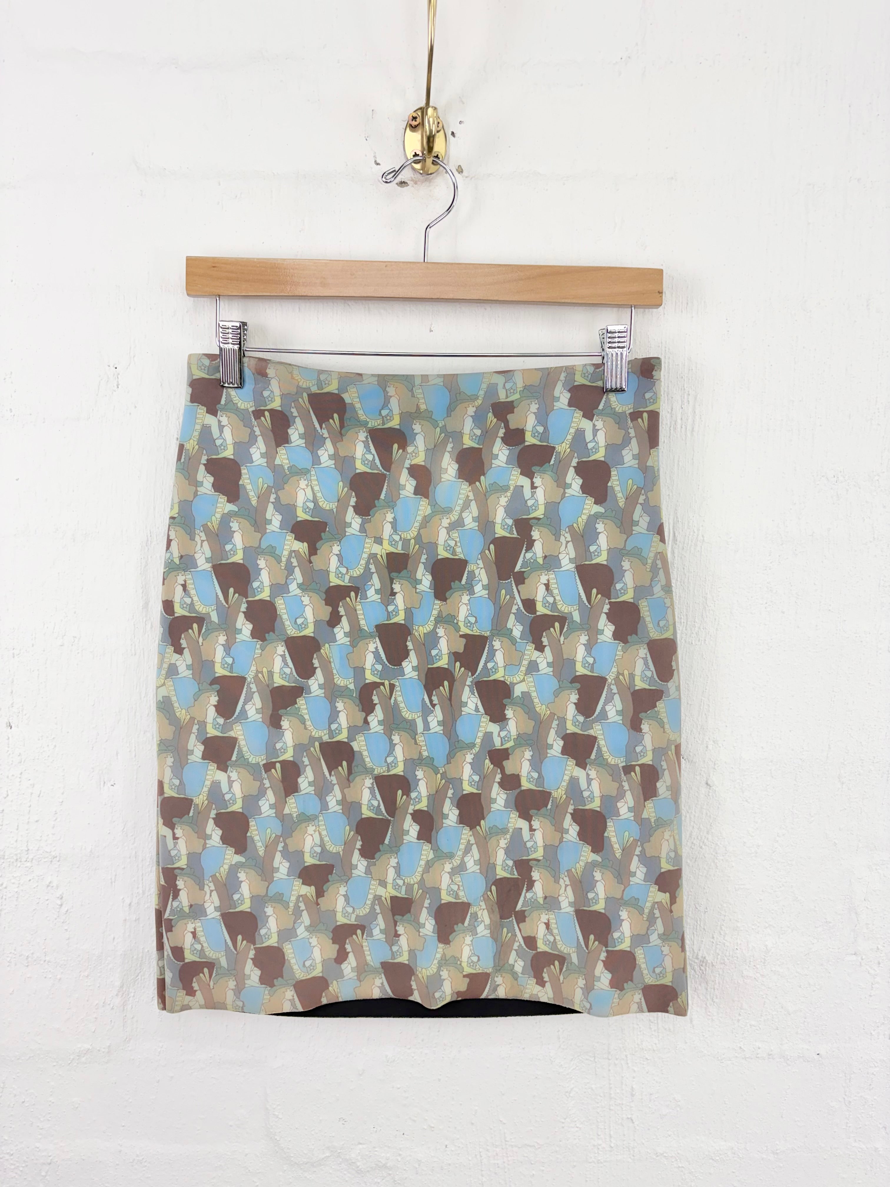 A patterned mini skirt with an abstract geometric print in brown, beige, and light blue, hanging from a wooden hanger against a white brick wall.
