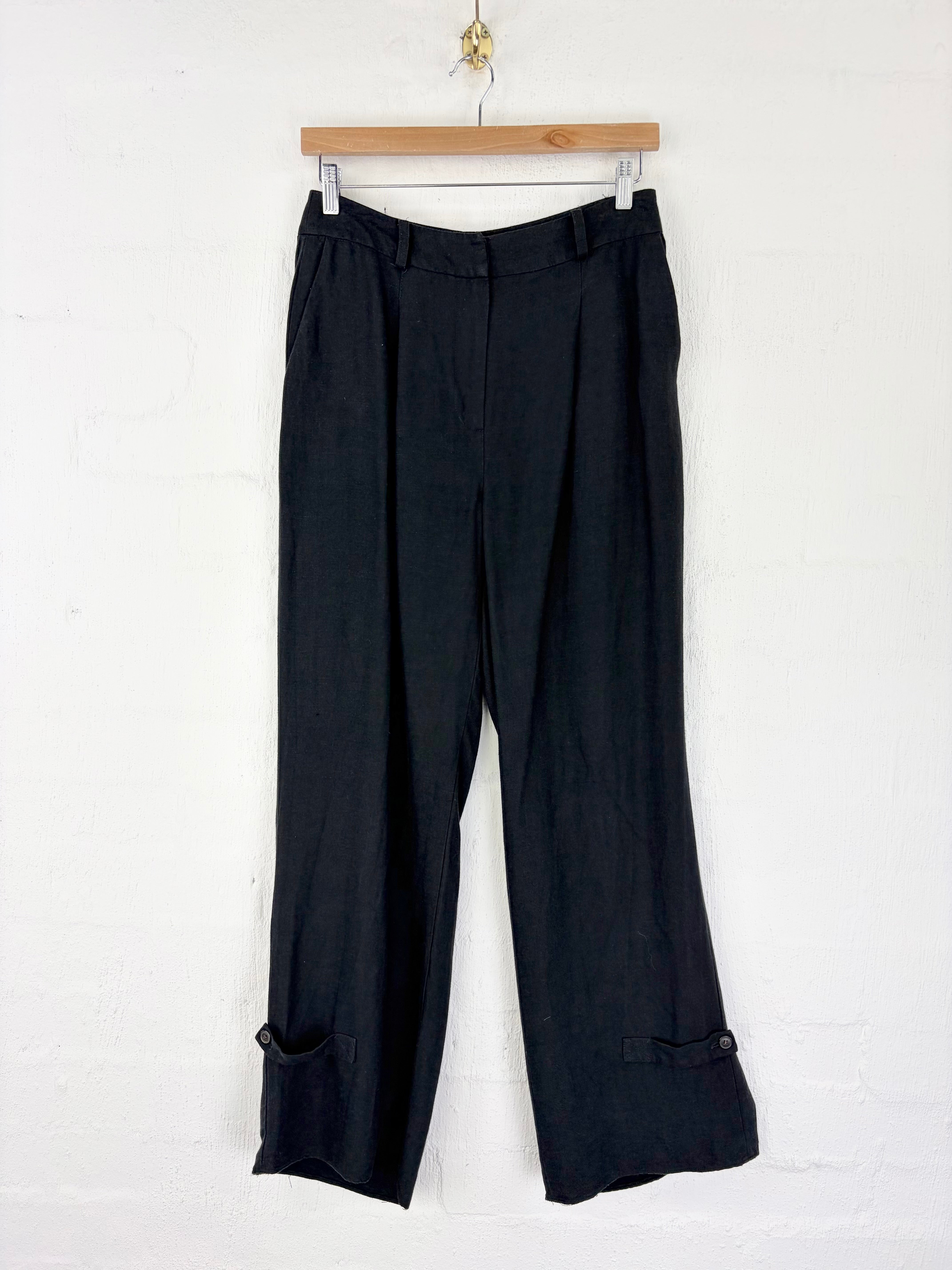 Black wide-leg cropped trousers with buttoned cuff details, hanging from a wooden hanger against a white painted brick wall.