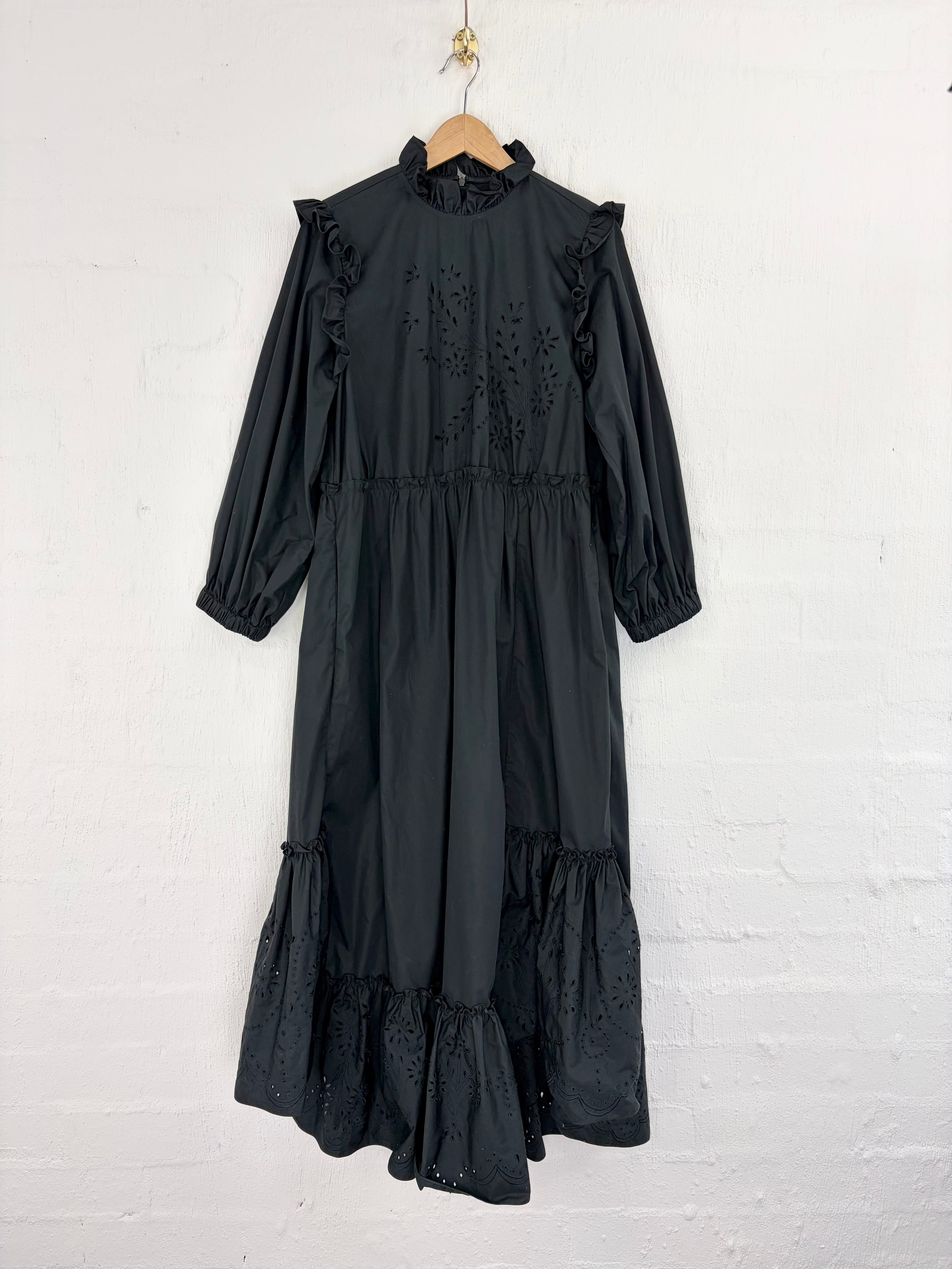 A black long-sleeve midi dress with eyelet embroidery and ruffle details, hanging on a wooden hanger against a white painted brick wall.