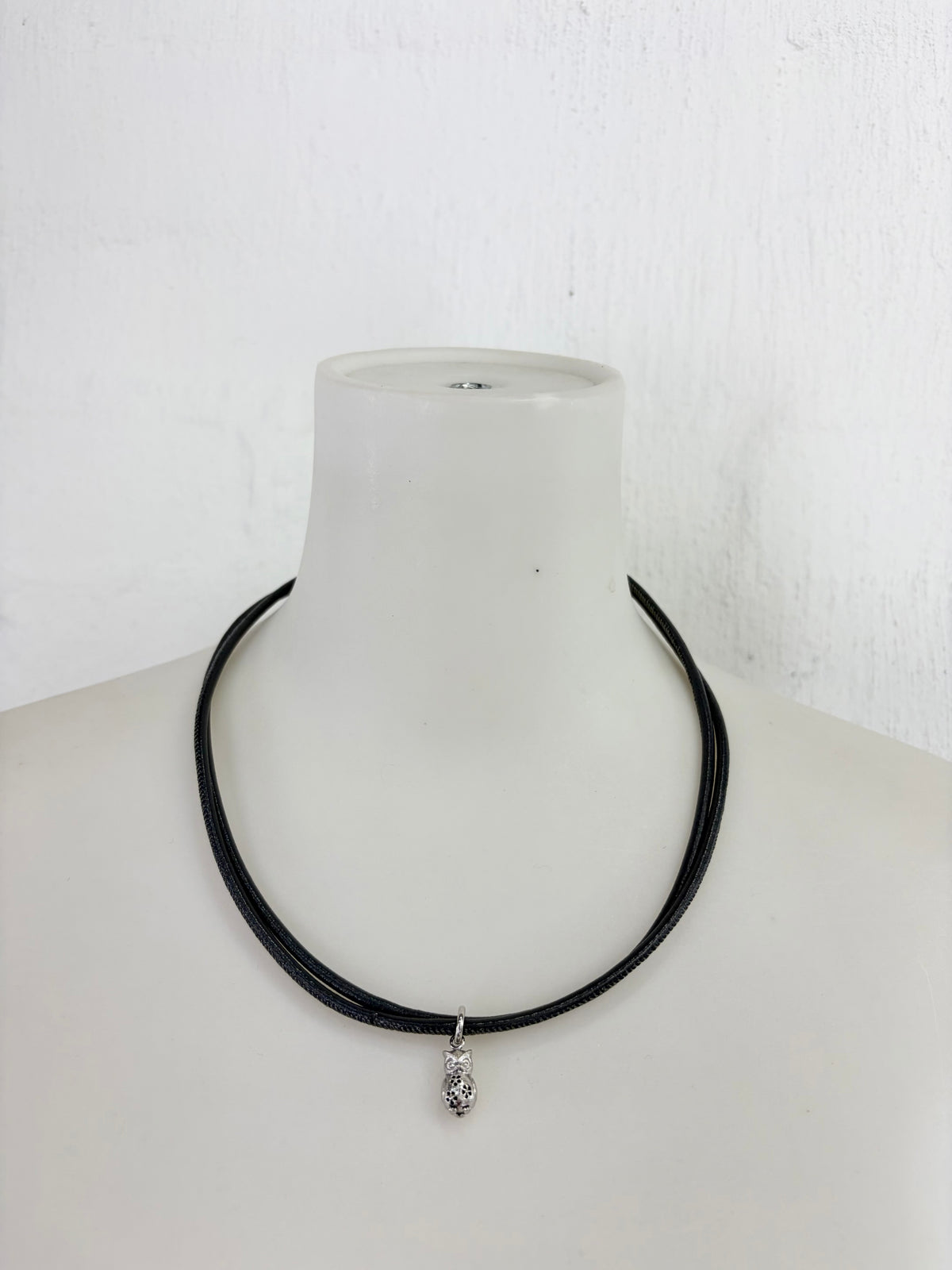 A black multi-strand cord necklace with a small silver teardrop pendant, displayed on a white mannequin bust against a white textured wall.